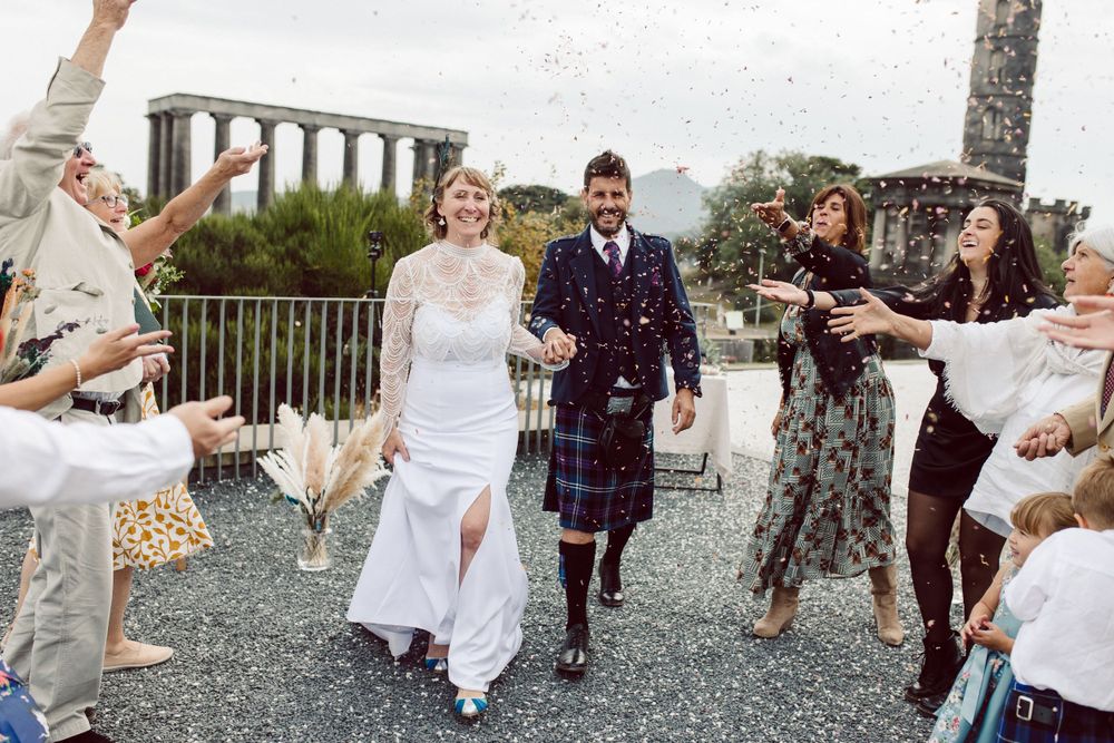 Wedding party throws confetti during outdoor celebration with stone columns in background.