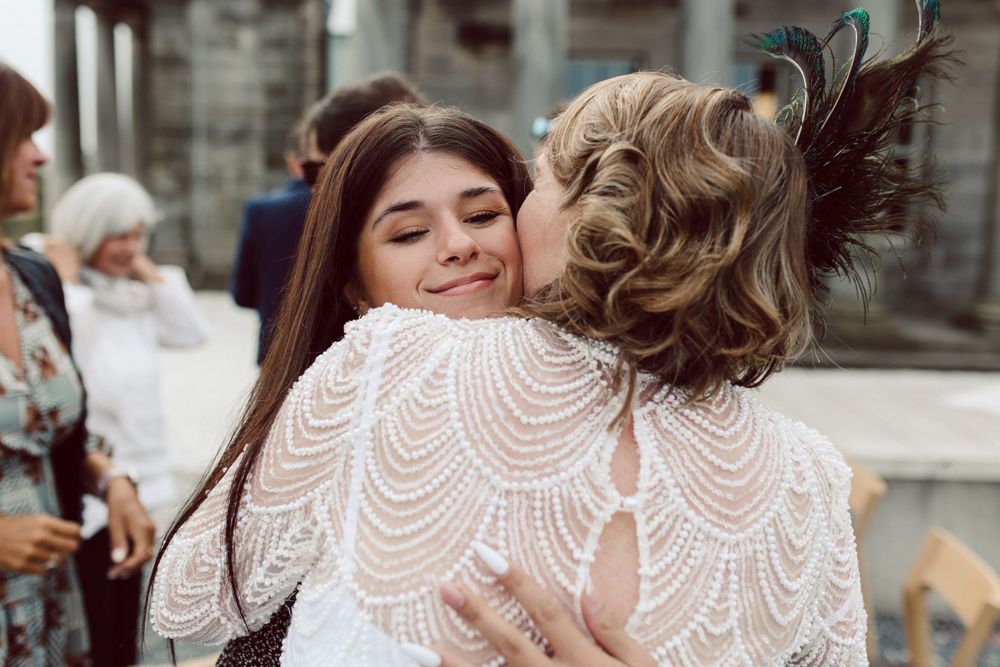 Wedding guests share emotional embraces during outdoor reception by stone architecture.