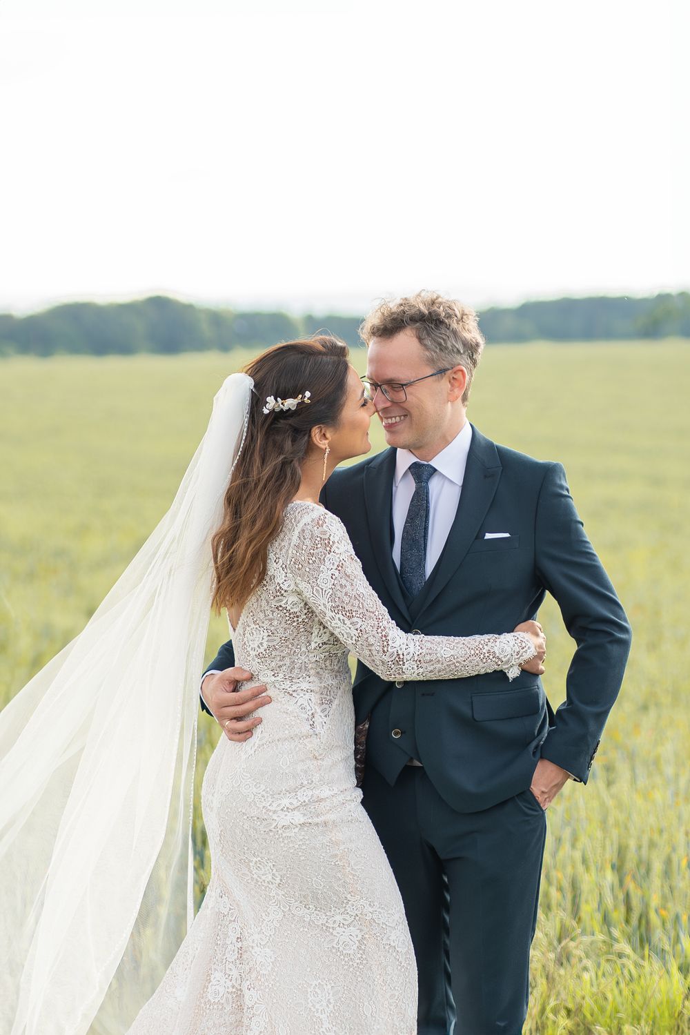 Romantic embrace between newlyweds in flowing veil against green field backdrop.