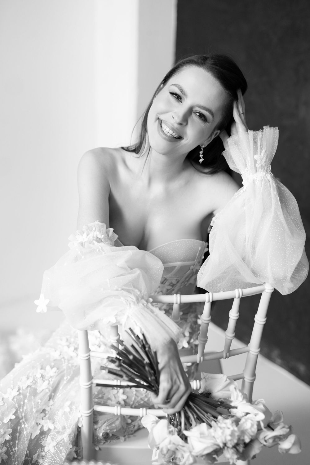 Black and white portrait of a bride in a romantic wedding dress sitting on a chair with a joyful expression.