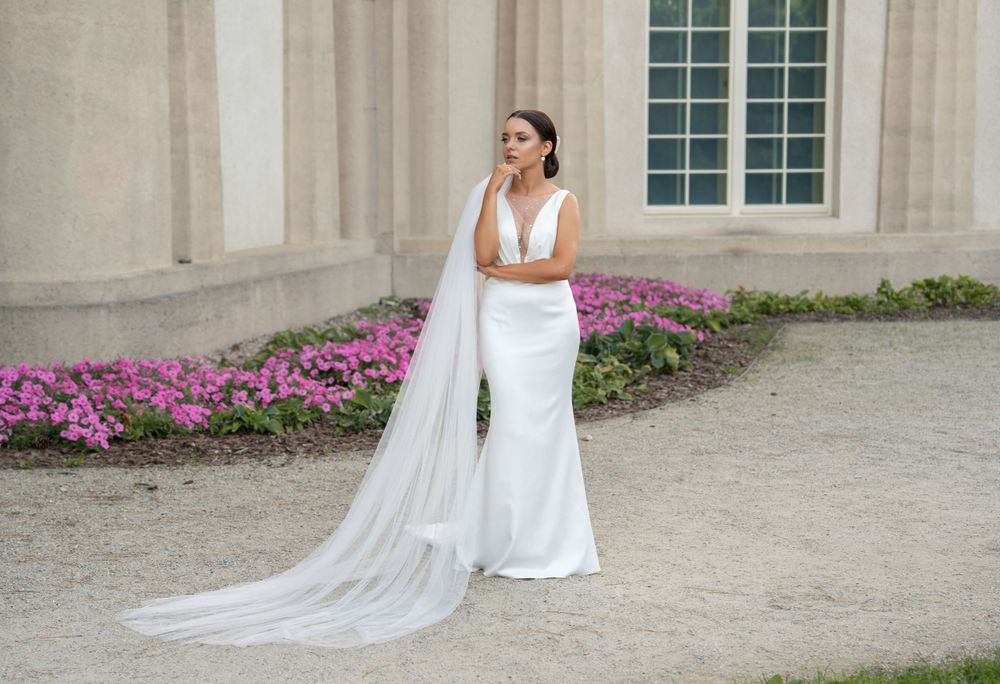 Elegant bride in fitted white dress and cathedral veil poses by stone building with purple flowers.
