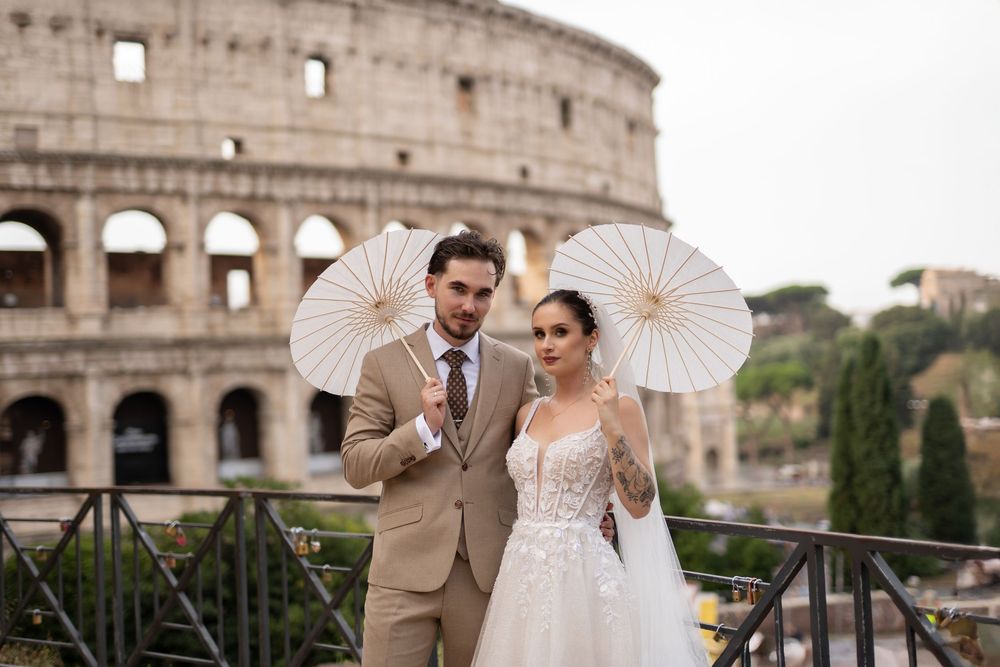 A couple holding white parasols poses in elegant wedding attire in front of the historic Roman Colosseum.