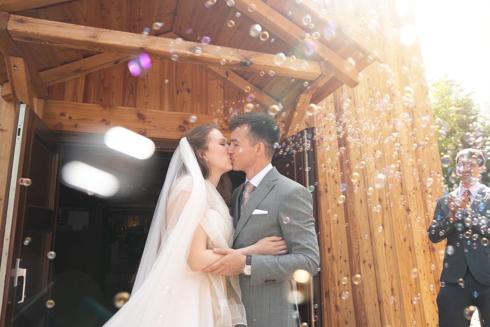 A bride and groom share a kiss at a wooden chapel entrance, surrounded by floating bubbles on their wedding day.