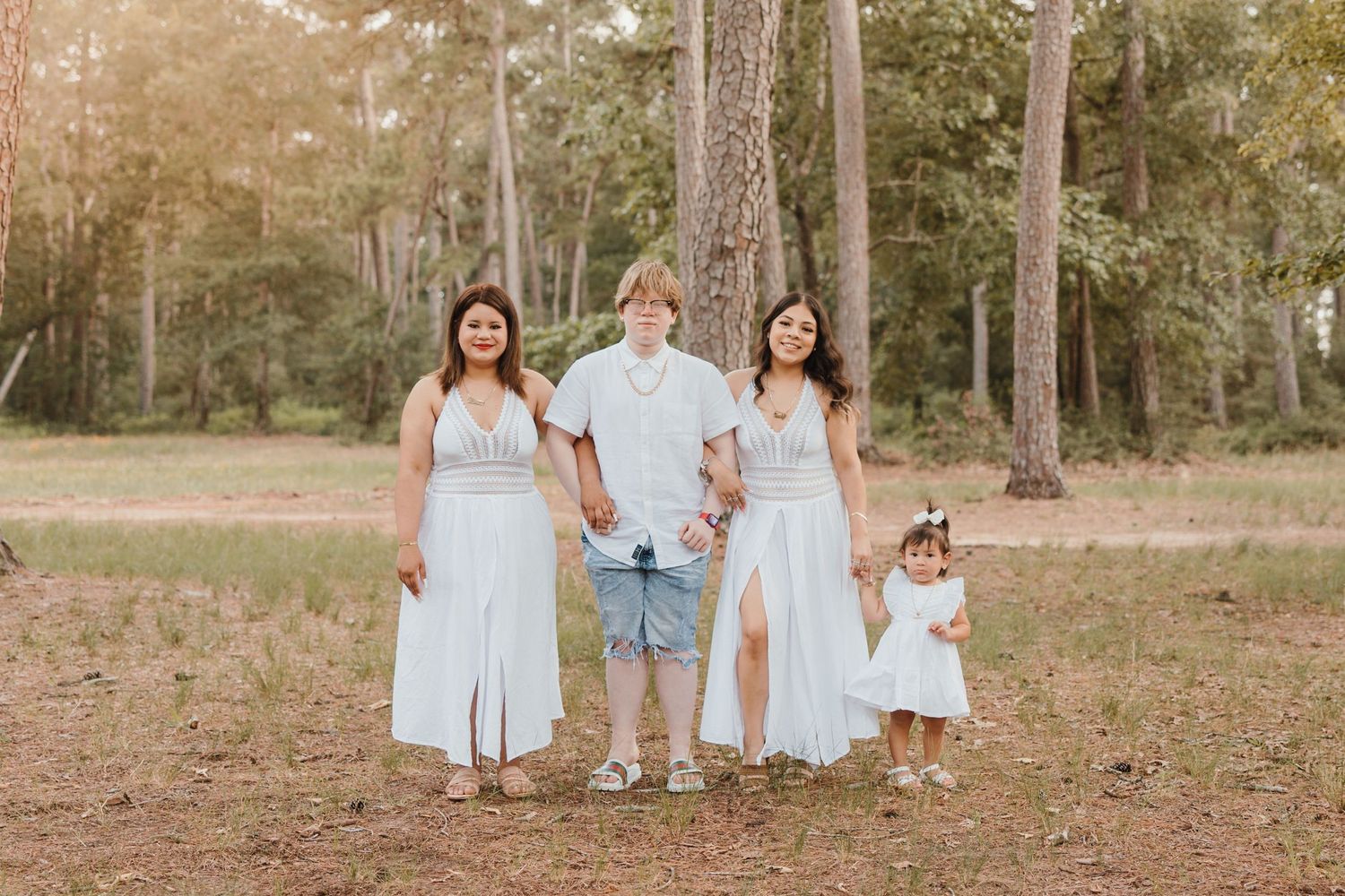 A family wearing coordinated white outfits poses together in a wooded outdoor setting at sunset.