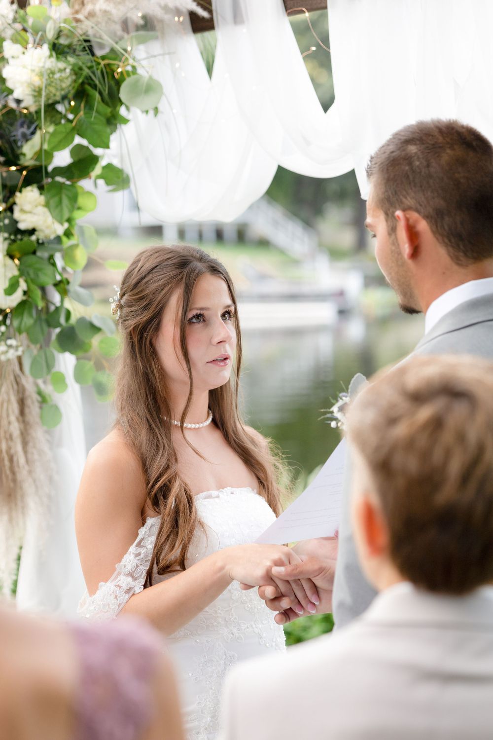 Outdoor wedding ceremony under white draped arch with greenery and lake backdrop.