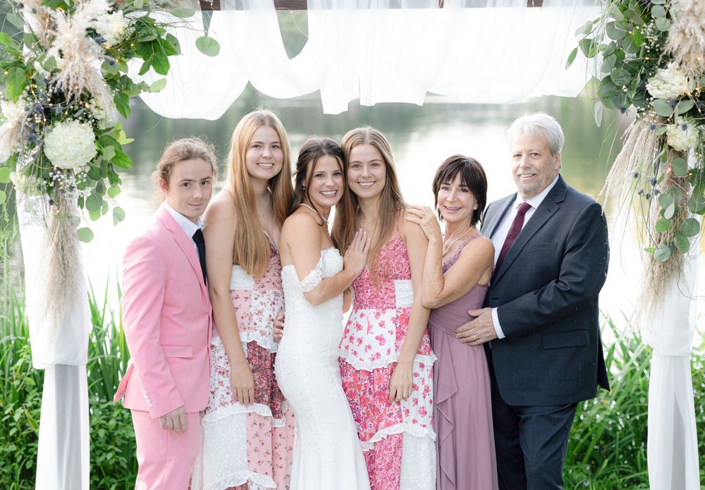 Wedding party in pink and white attire poses under decorated outdoor arch.
