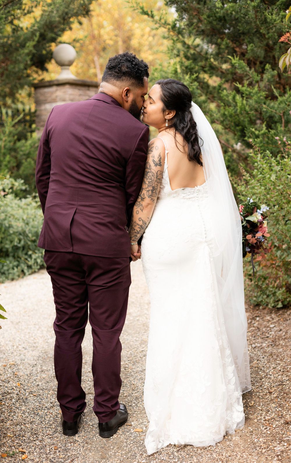 Couple in burgundy suit and white wedding dress walking on garden path.