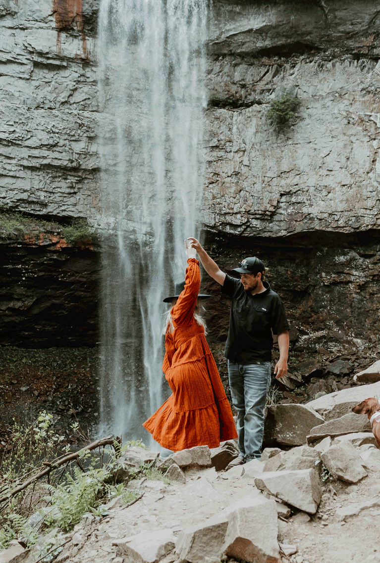 Couple explores rocky terrain near dramatic waterfall, woman wearing vibrant orange dress.