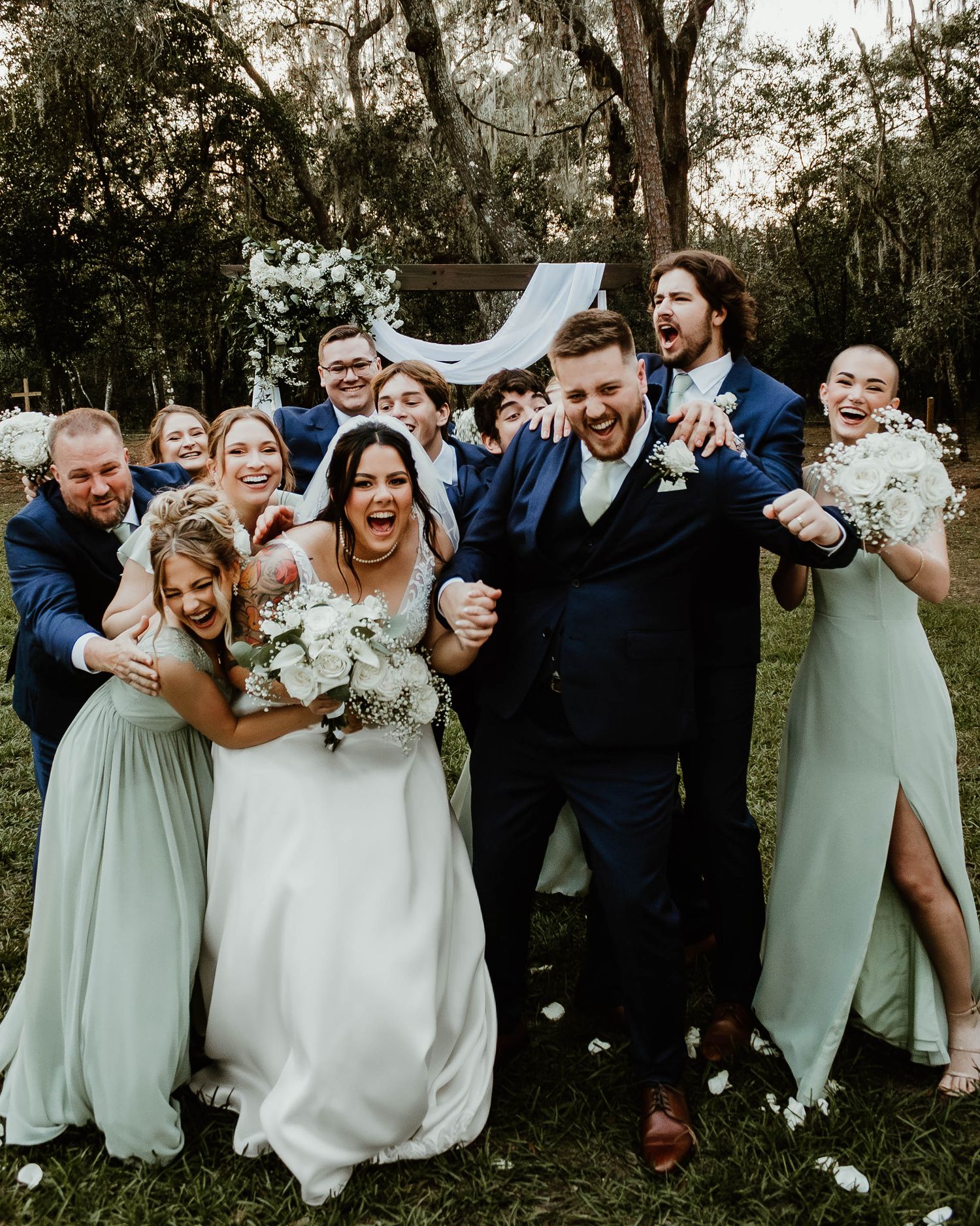 An energetic wedding party poses for fun group photos in a garden setting.