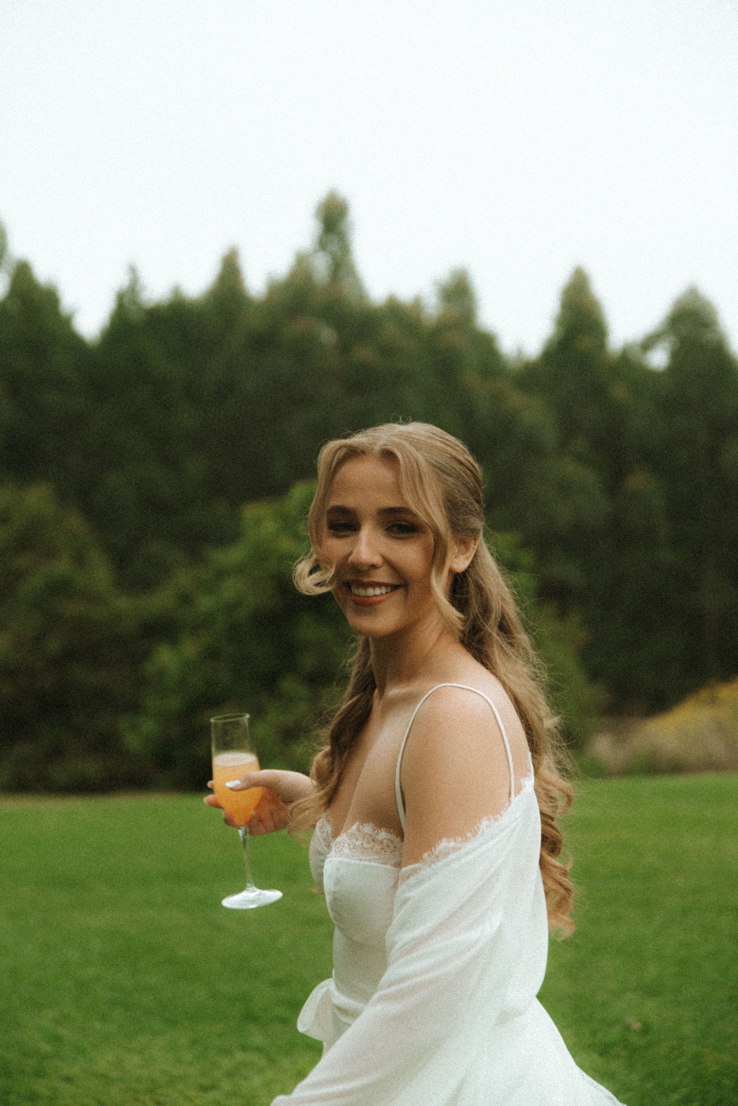 A person in a white dress twirls joyfully while holding a champagne glass in a green outdoor field.
