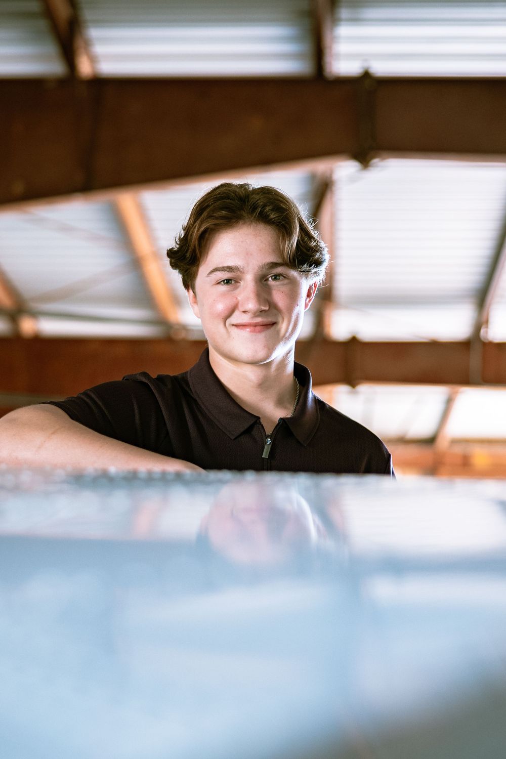 A person in a black polo shirt smiling warmly indoors near a window with natural light streaming in.