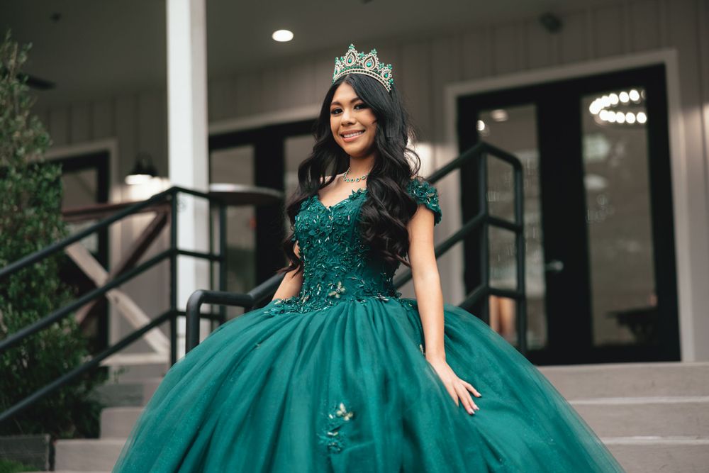 A woman wearing a sparkling emerald green ball gown and crown stands elegantly on a staircase at night.