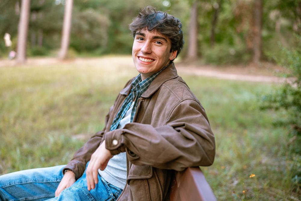 Someone wearing a brown jacket and hat smiles while sitting outdoors in a natural setting.