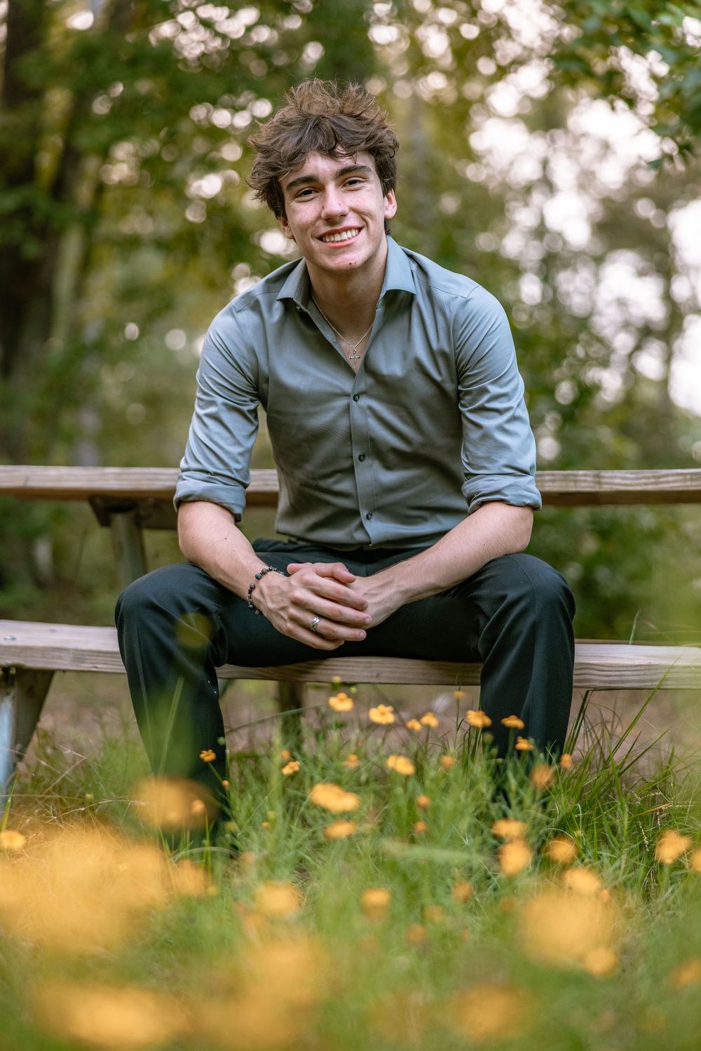 Portrait of someone sitting on a wooden bench outdoors with yellow wildflowers in the foreground and trees in the background.