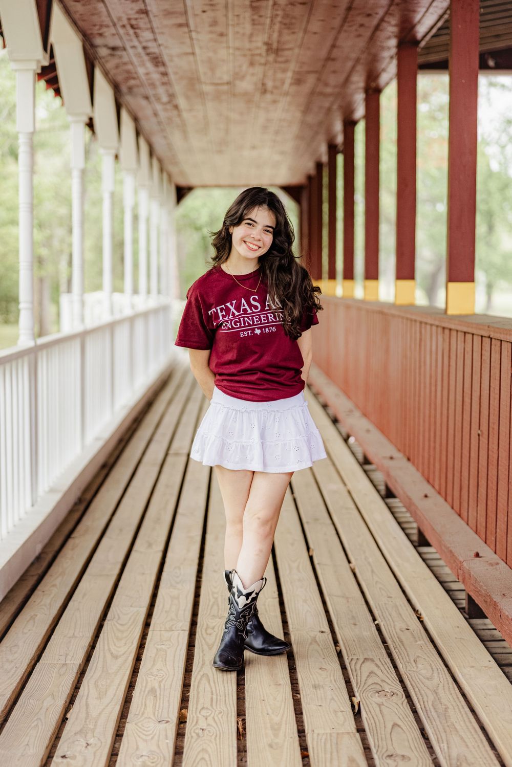 A student in a maroon Texas A&M t-shirt, white skirt and black boots stands in a covered wooden walkway.