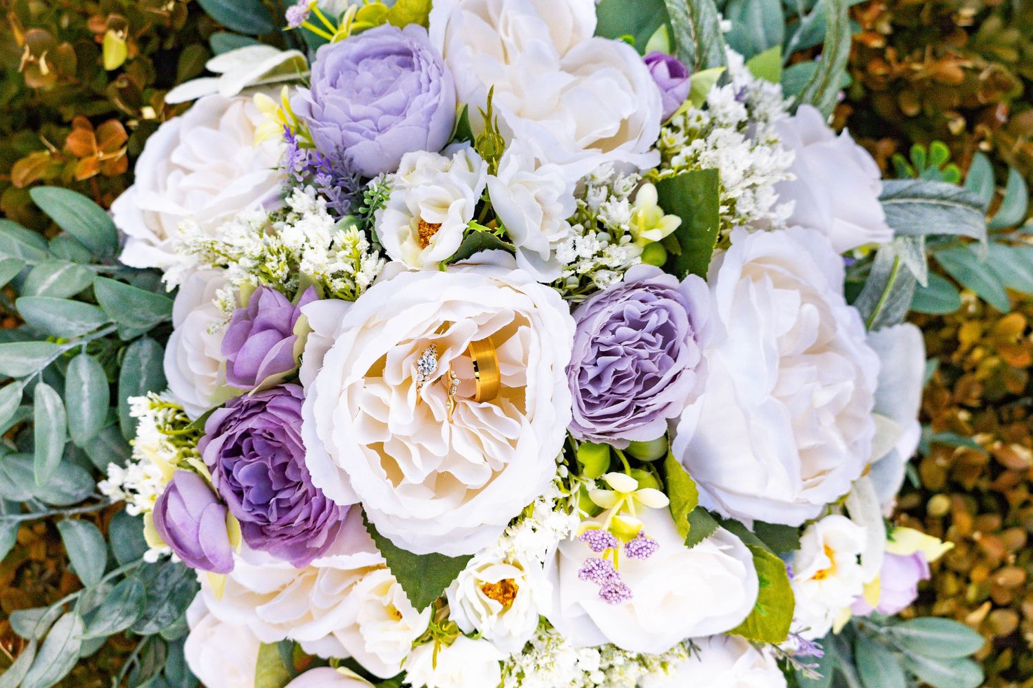 A lush wedding bouquet featuring purple and white roses with delicate greenery and small white accent flowers with golden rings.