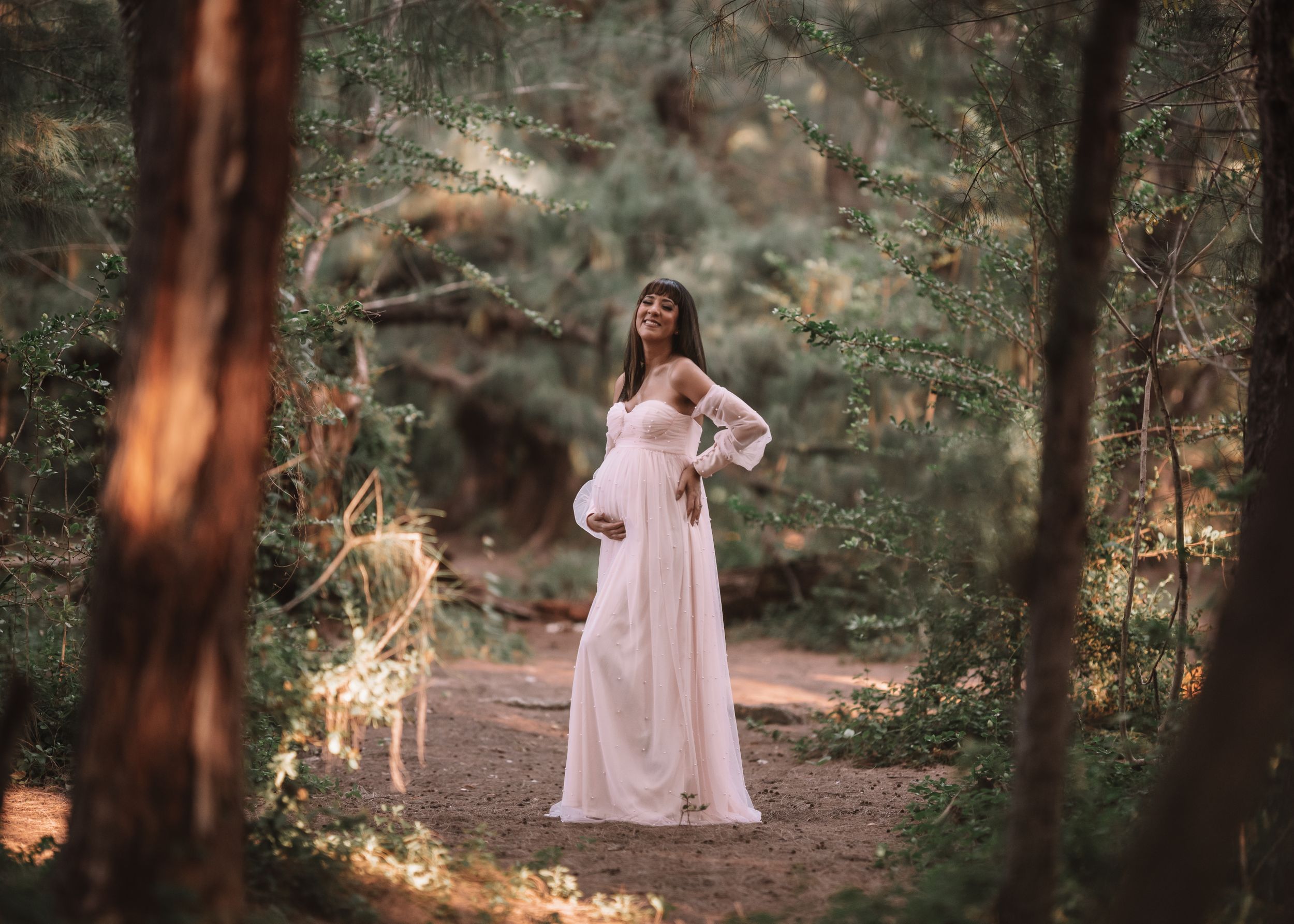 A woman in a flowing pink dress poses gracefully in a sunlit forest setting.