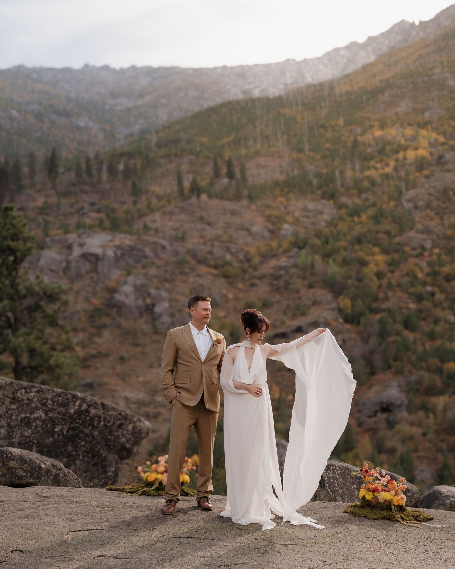 A couple poses together on a rocky mountain ledge with dramatic desert landscape views at sunset.