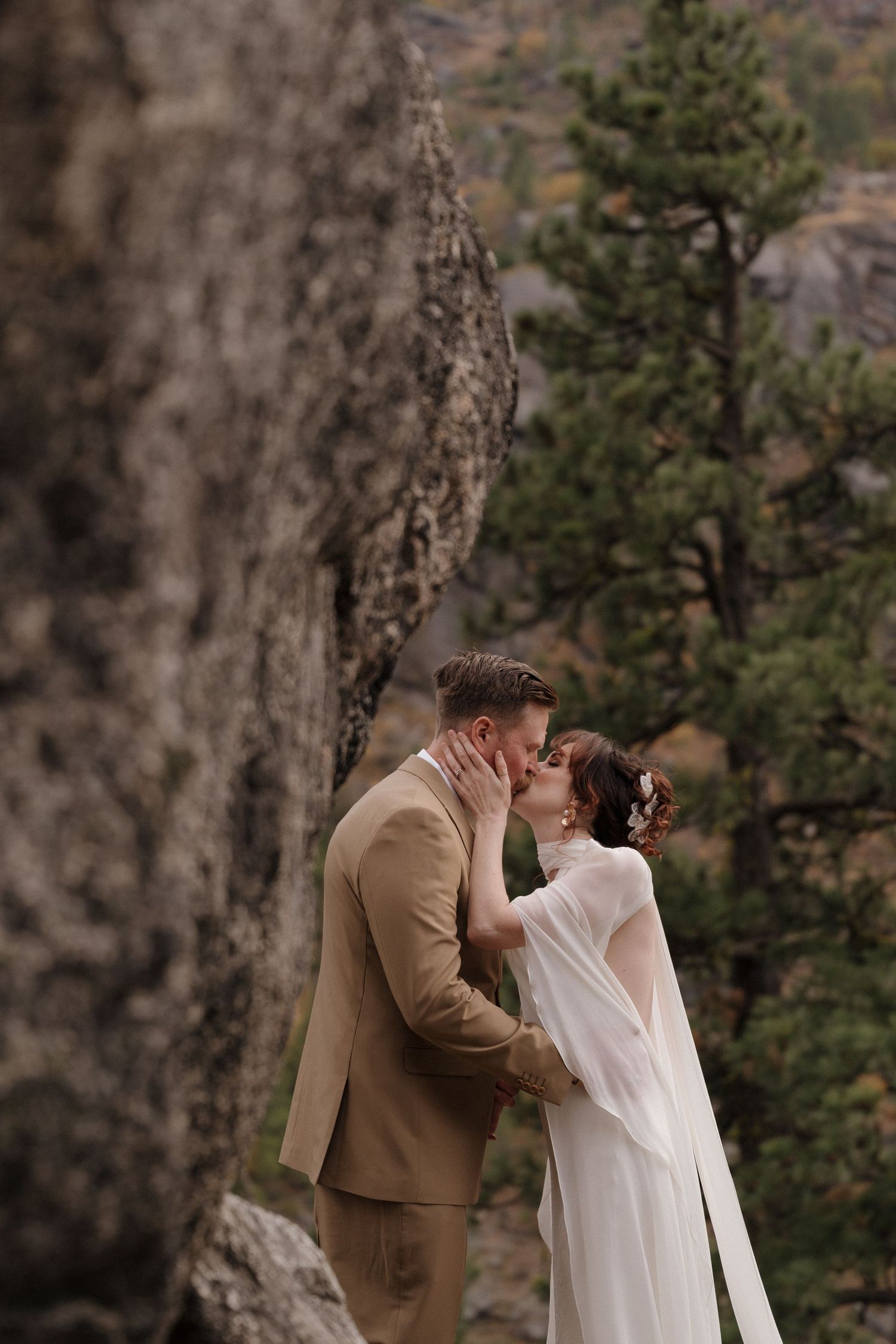 A couple shares a romantic kiss outdoors against a rocky mountainous backdrop with evergreen trees.