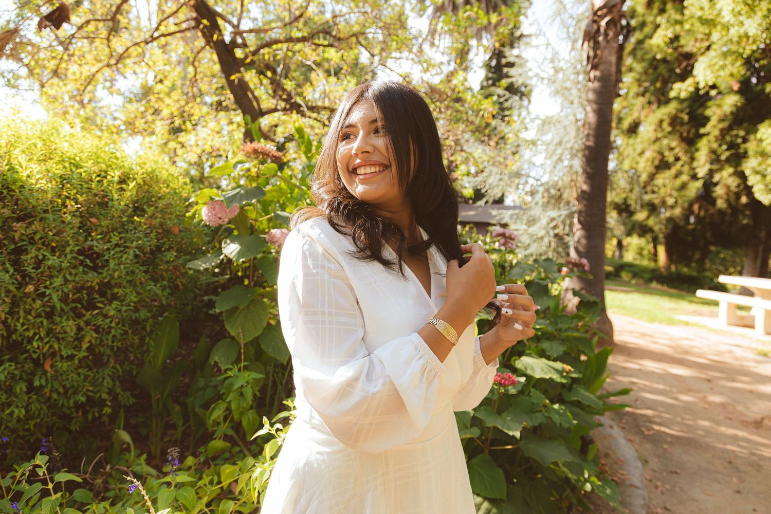 A woman in white dress poses joyfully in a sunny garden setting.