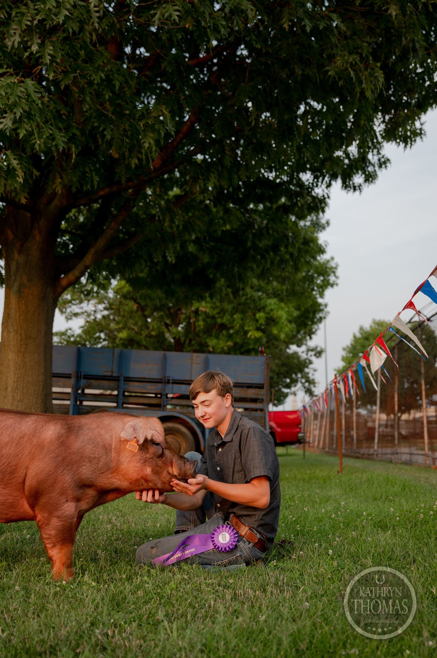 A young exhibitor kneels beside a reddish-brown show pig in a grassy outdoor area with flags visible in the background.