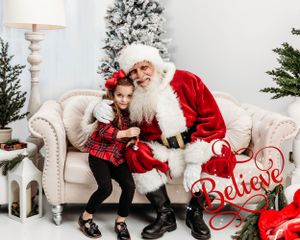A festive holiday scene with a white Christmas tree and red 'Believe' sign decoration. Santa photographer