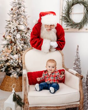 Holiday portraits on cream chair with flocked Christmas tree and wreath in background. Santa photographer