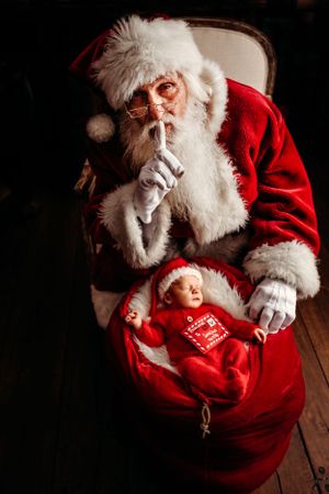 A dimly lit Christmas scene with traditional red and white holiday attire and a gift sack with newborn. Santa photographer