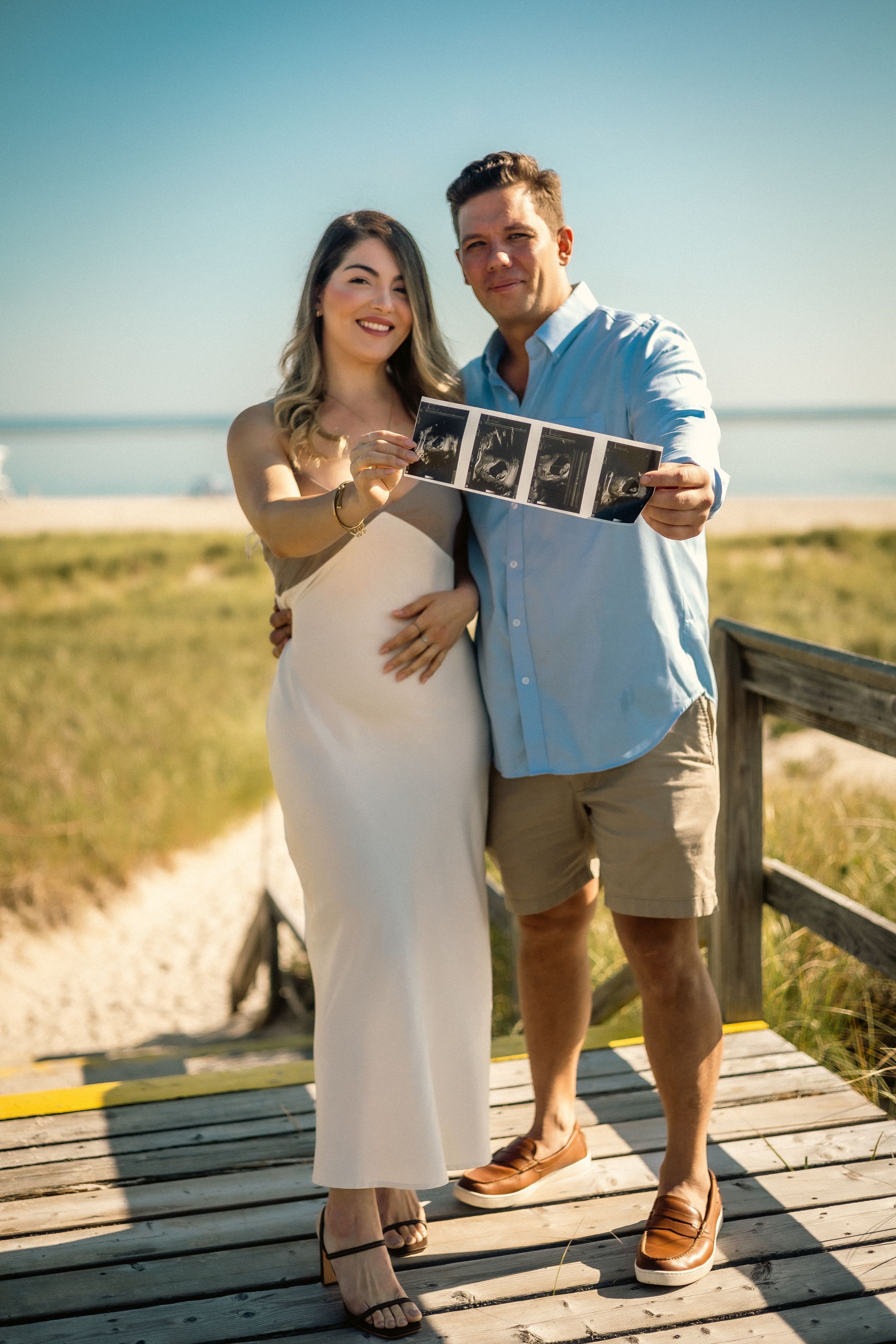 Couple on beach boardwalk holding ultrasound photos in sunny weather.