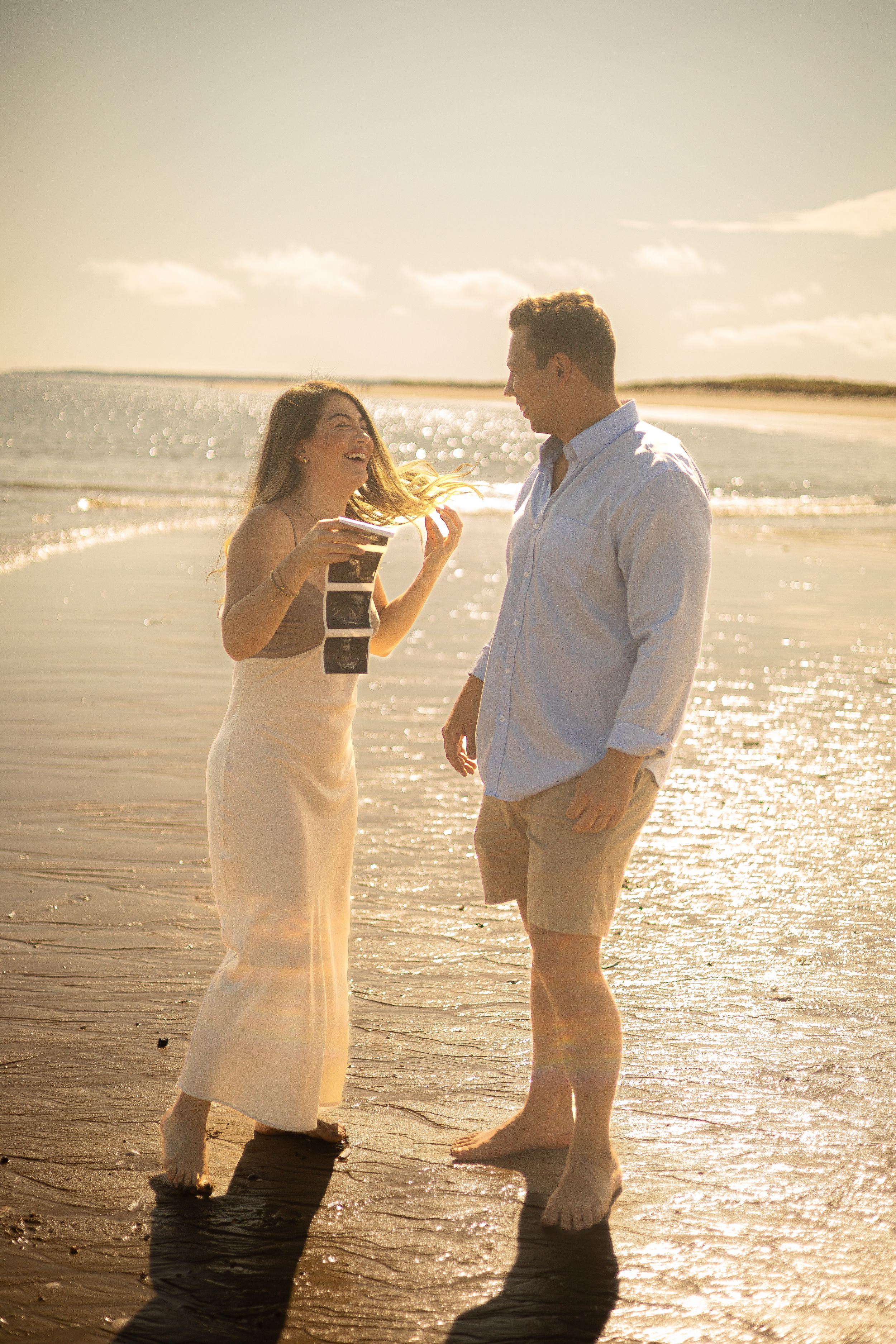 Couple sharing joyful moment on beach with ultrasound photos at sunset.