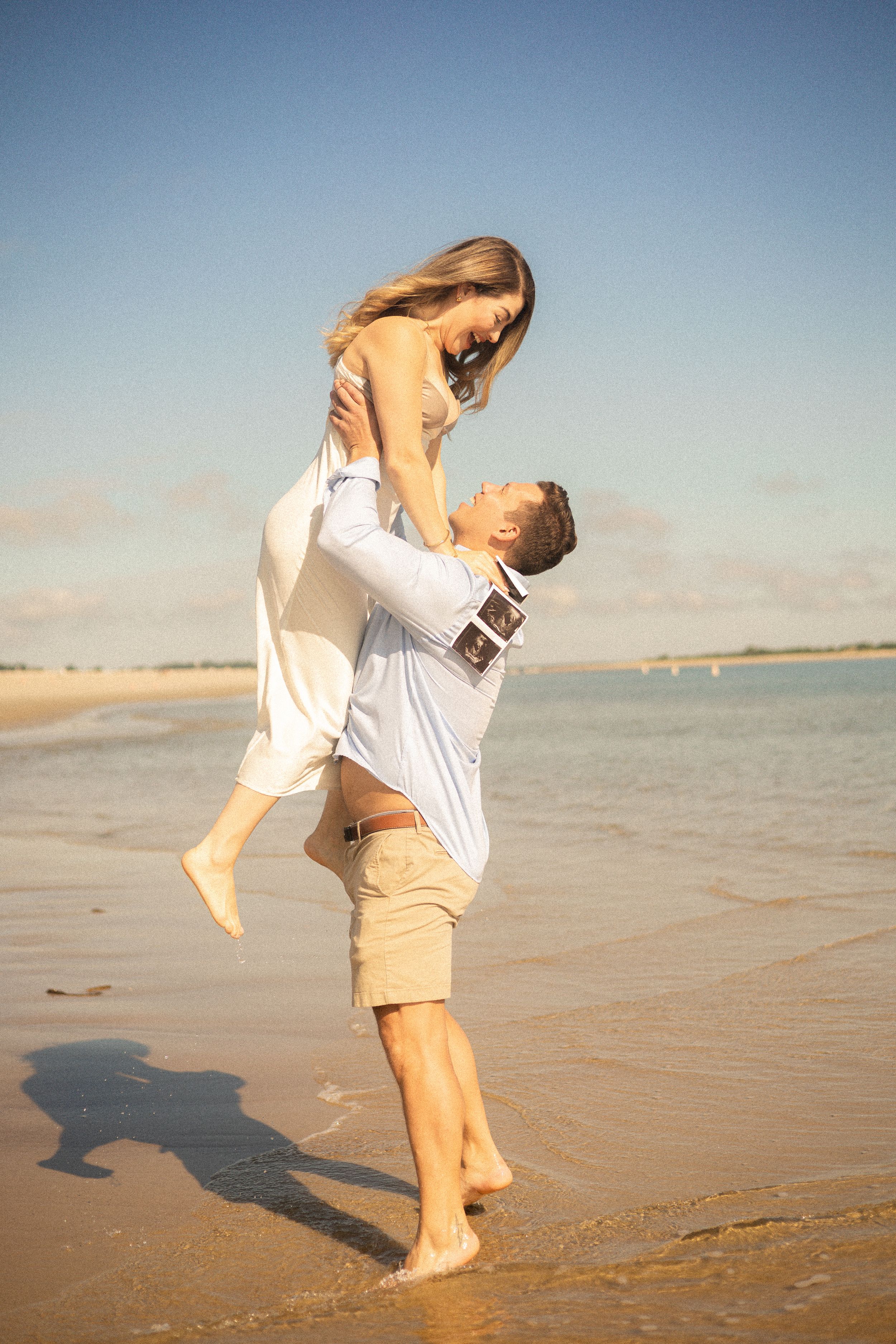 A romantic couple embraces on a sunny beach shoreline during sunset with waves in the background.