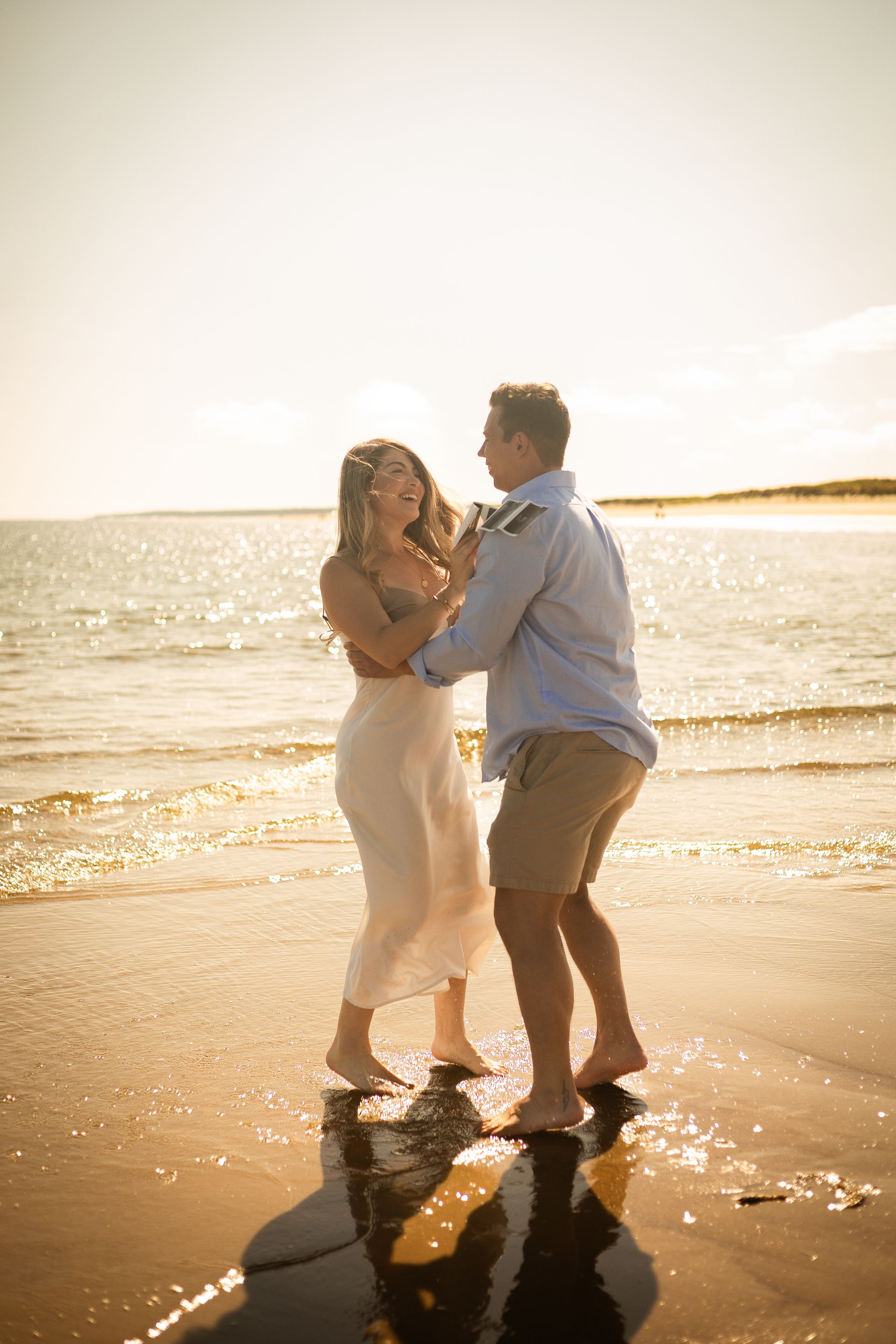 A couple dances together on a beach at golden hour with sunlit water behind them.