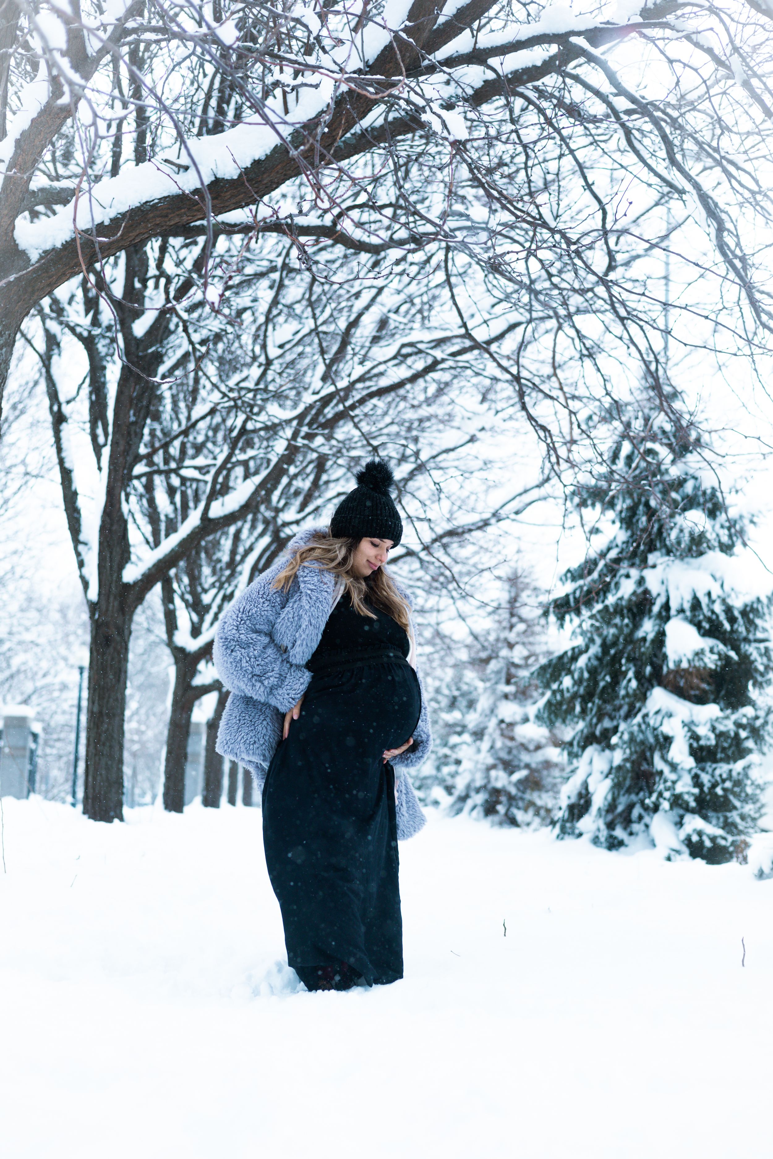 Winter maternity photoshoot in a snowy forest with a couple embracing in warm coats.
