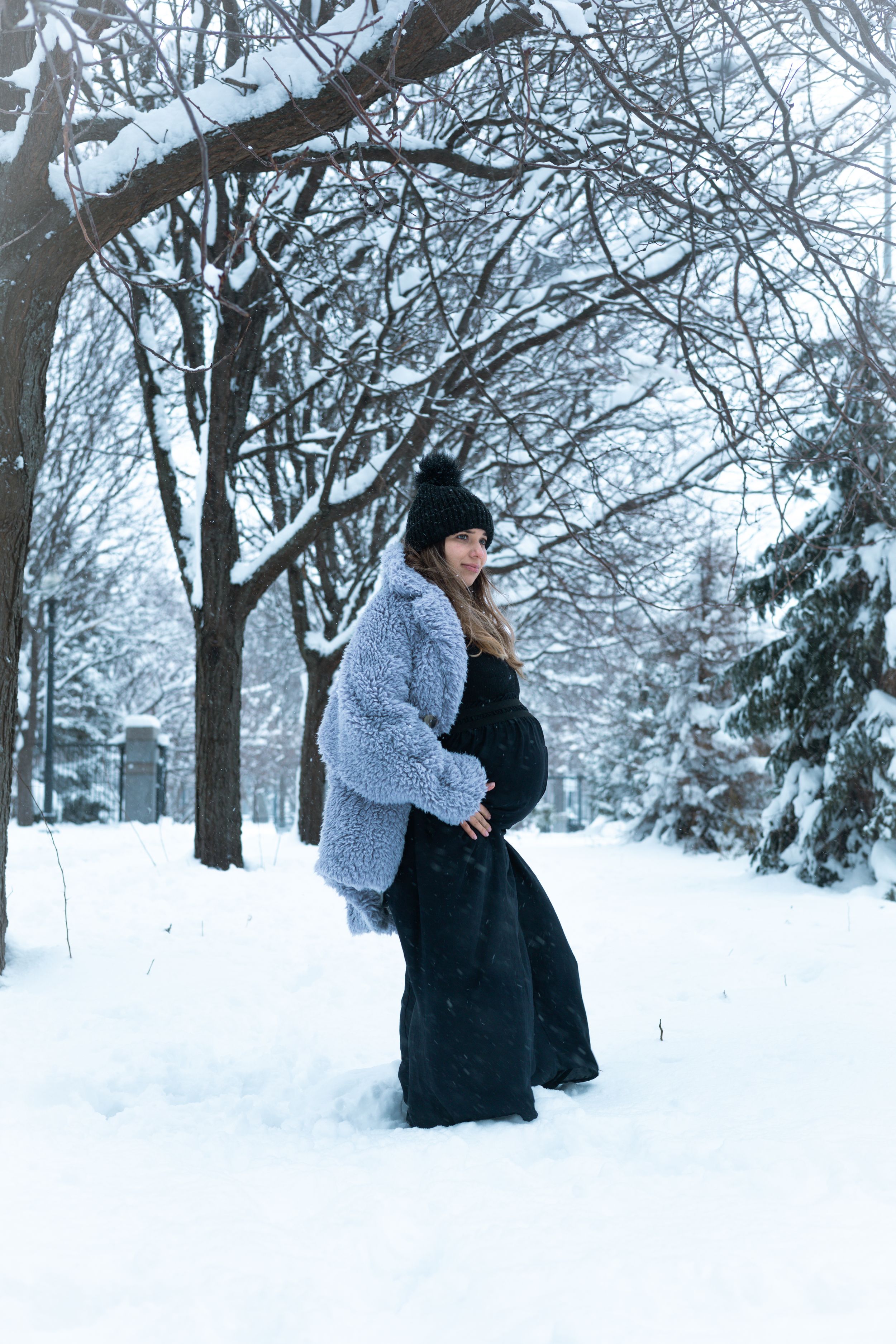 Expectant parent in a long dress and cozy cardigan poses in a snow-covered winter landscape.