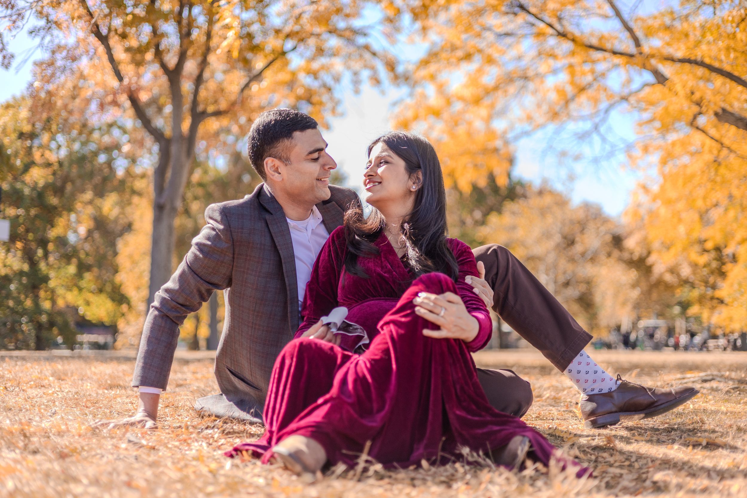 A couple sits together on fallen autumn leaves while sharing a joyful moment in a park with golden foliage.