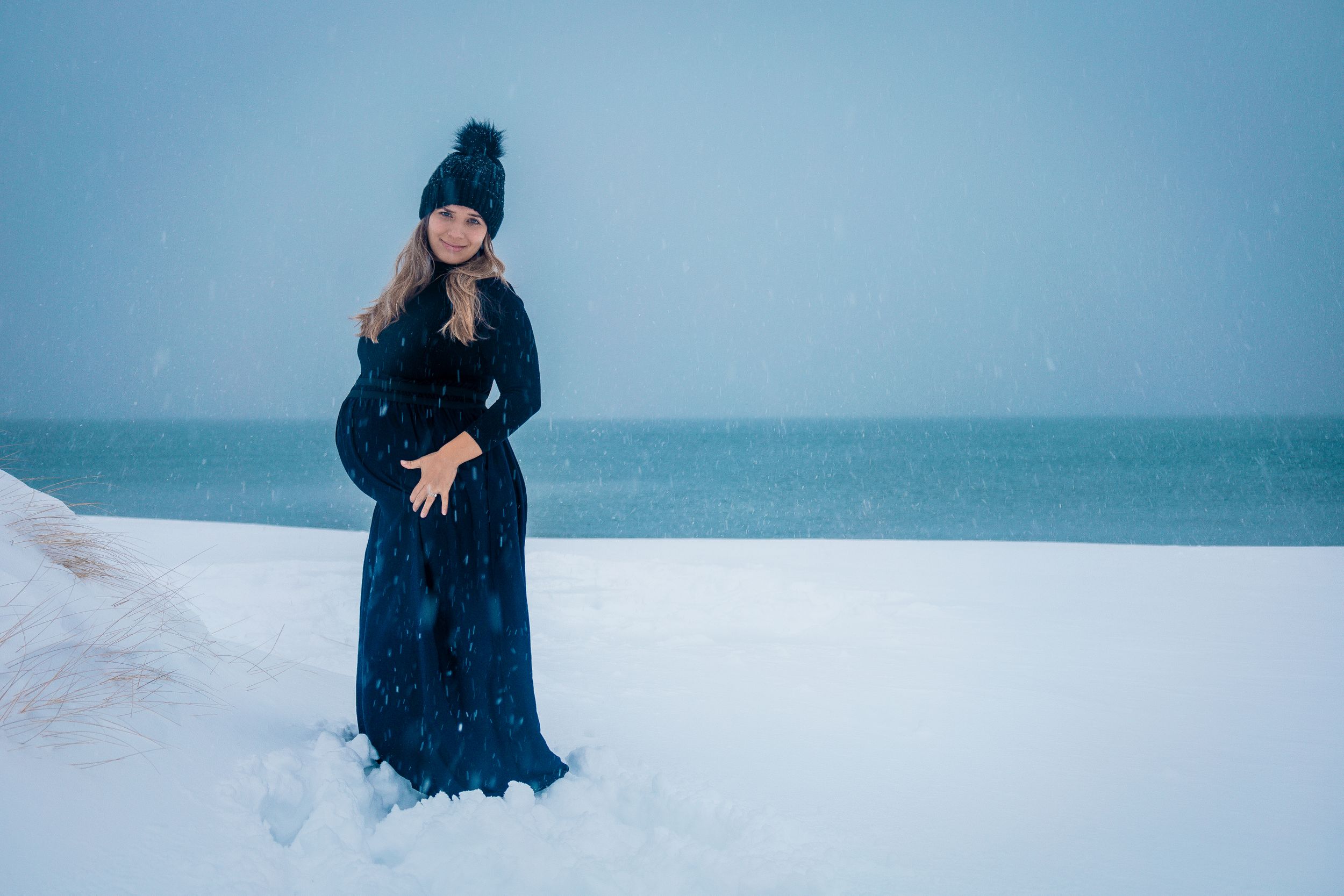 Figure in a dark dress and winter hat standing in a snowy landscape against a misty ocean backdrop.
