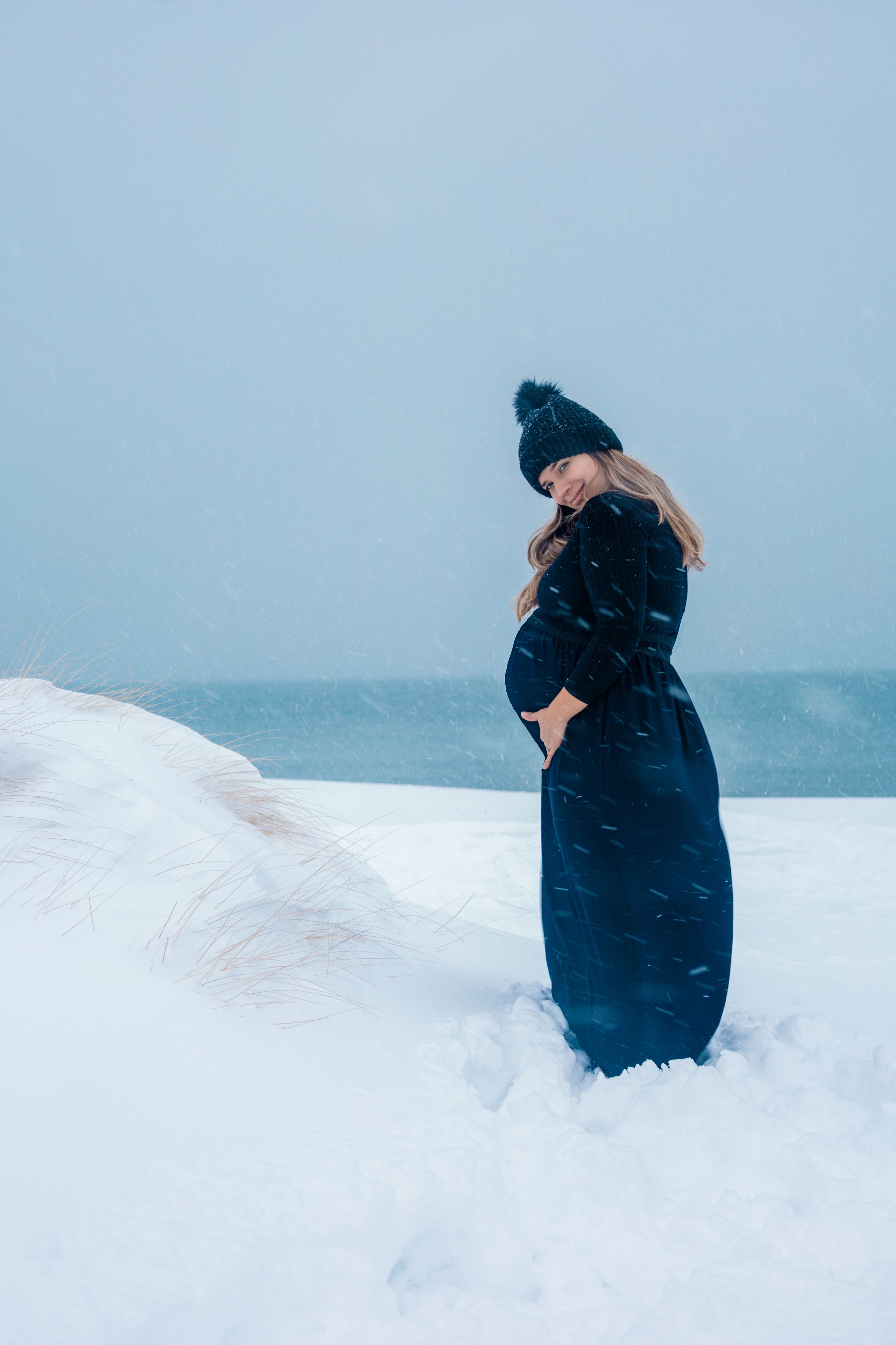 Maternity portrait of someone in a navy dress standing in deep snow against a misty backdrop.