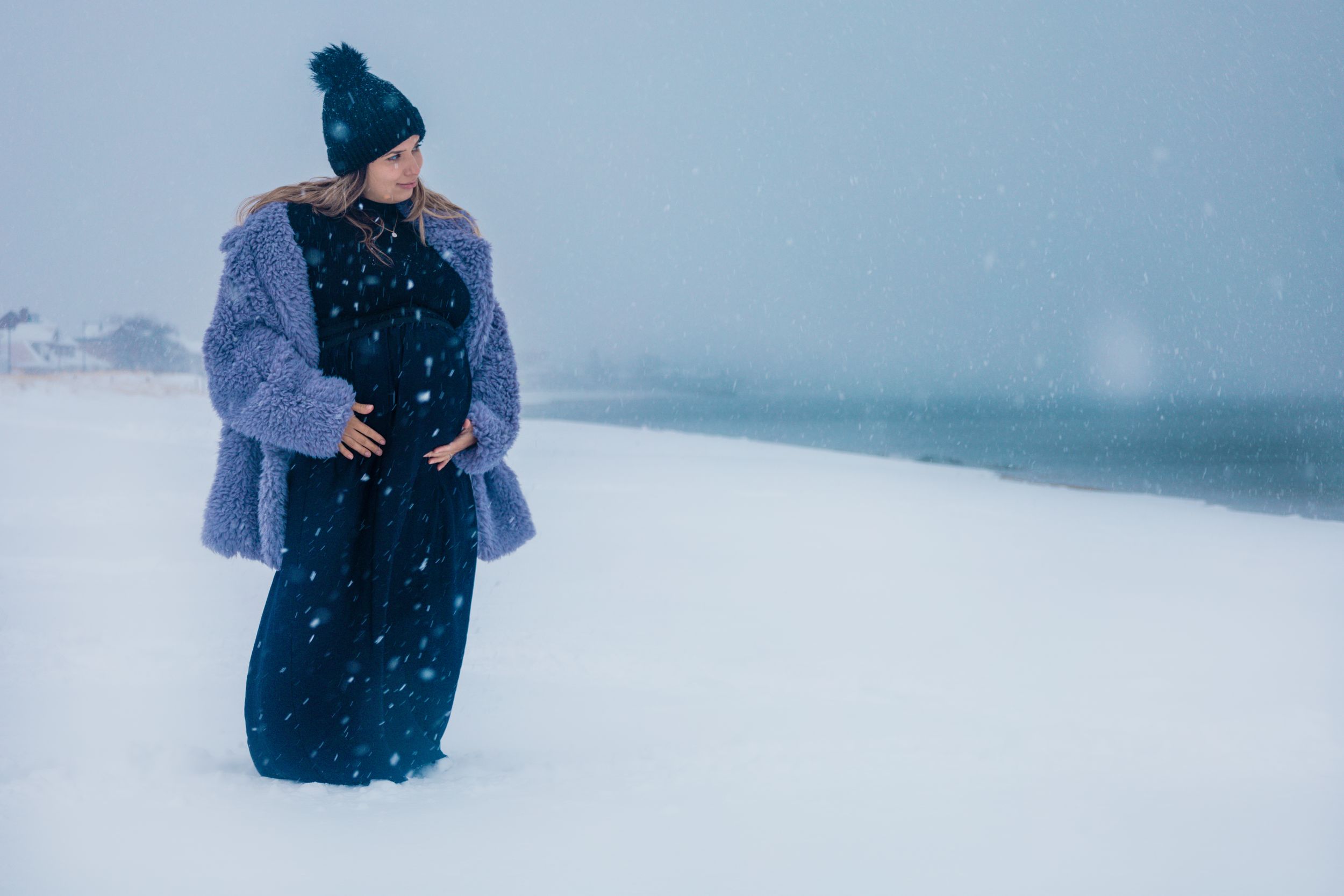 Person wearing winter clothing poses in a snowy coastal landscape with gray skies.