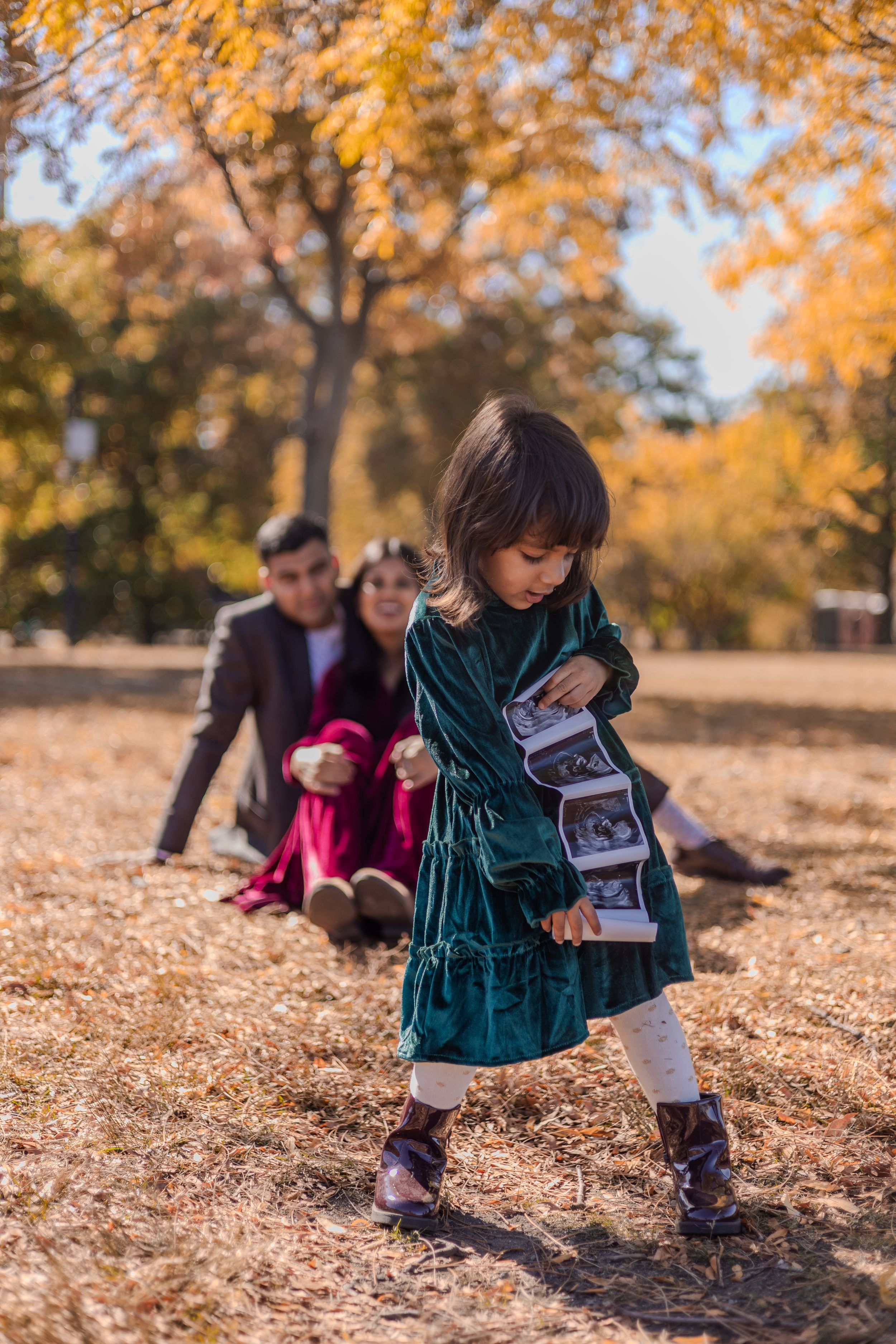 Child in green velvet dress looks at photos while standing on fallen autumn leaves in the park.