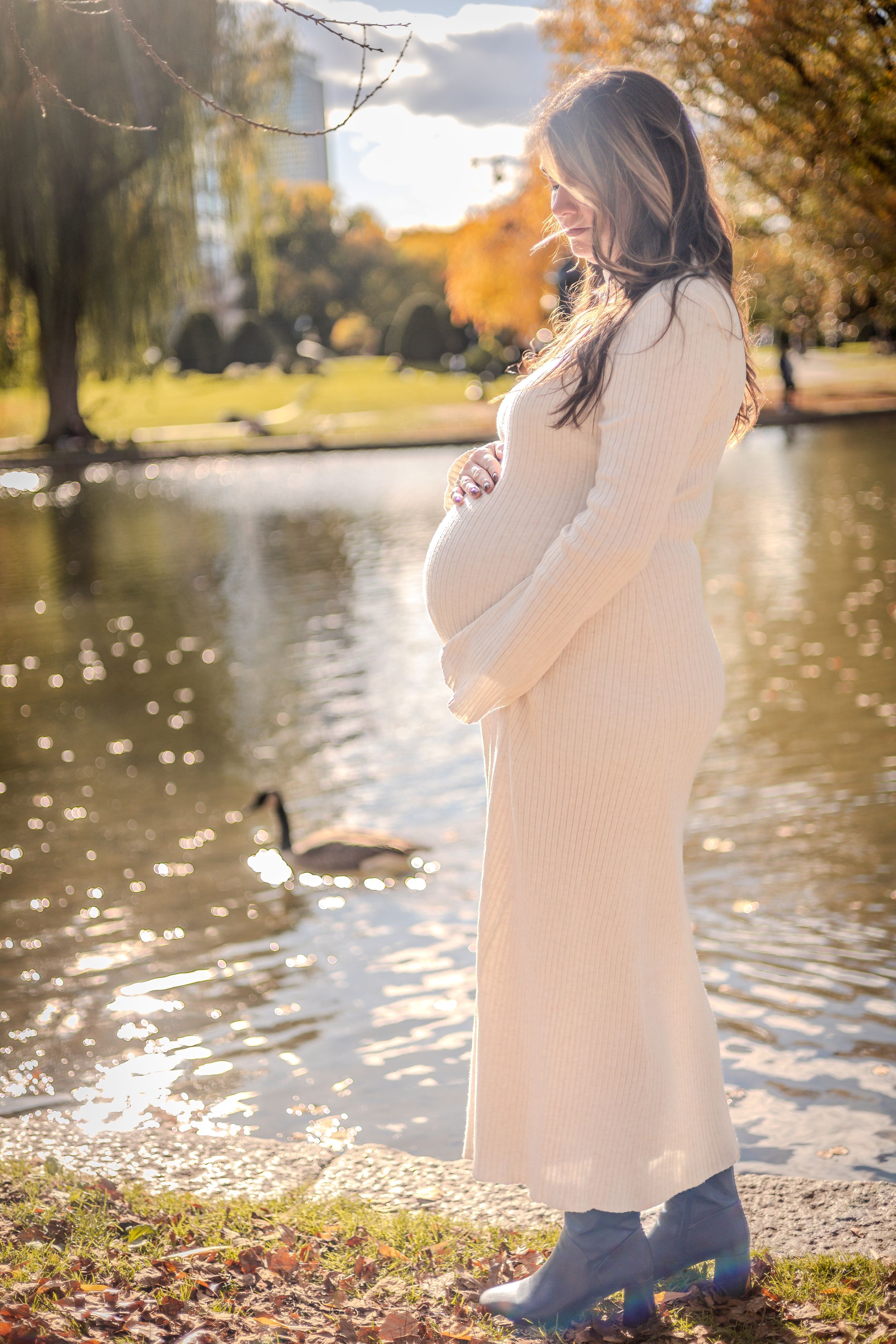 A pregnant figure in a cream sweater dress stands by a pond during autumn sunrise as a duck swims nearby.