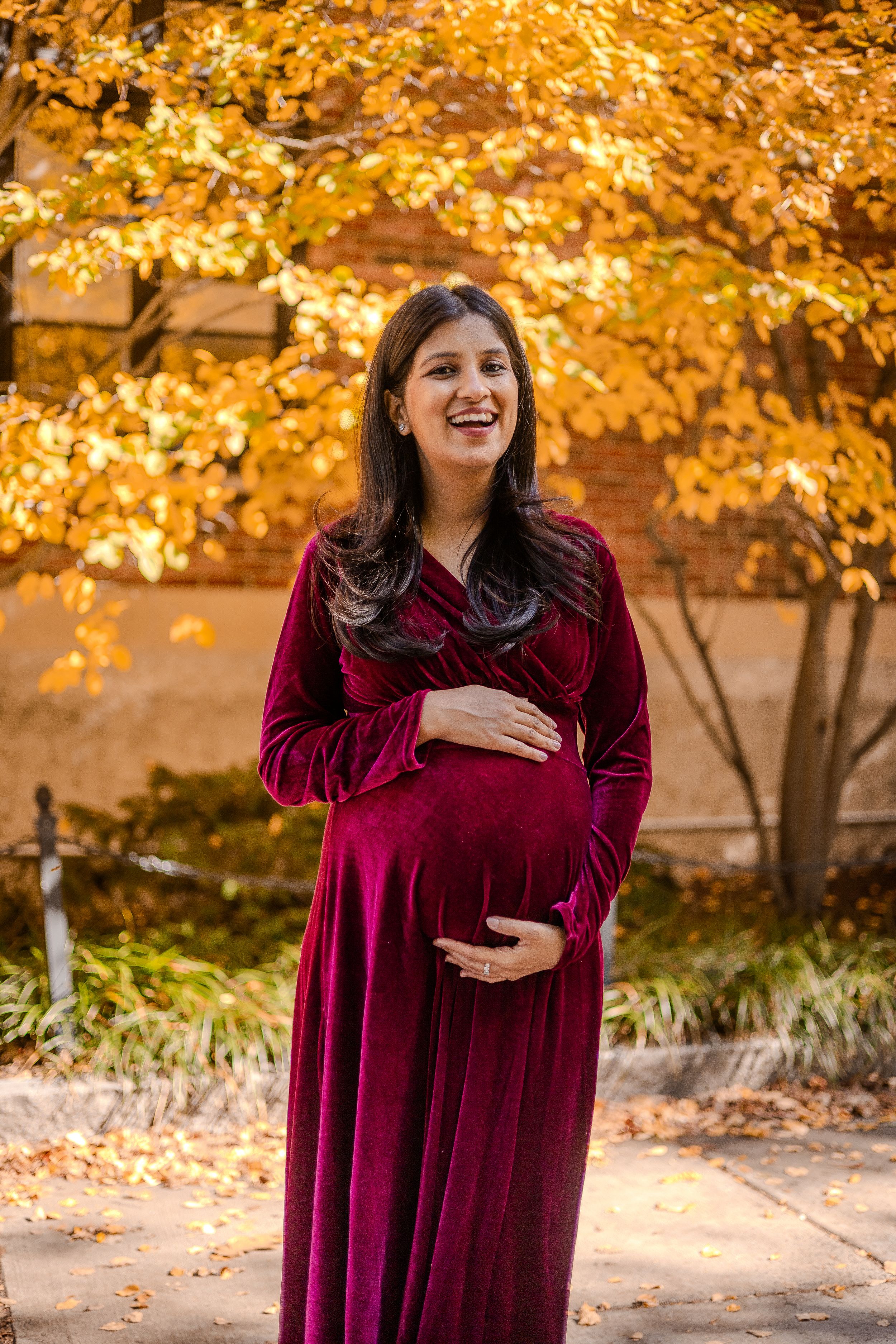 A pregnant person in a burgundy velvet dress stands outdoors with golden autumn foliage in the background.