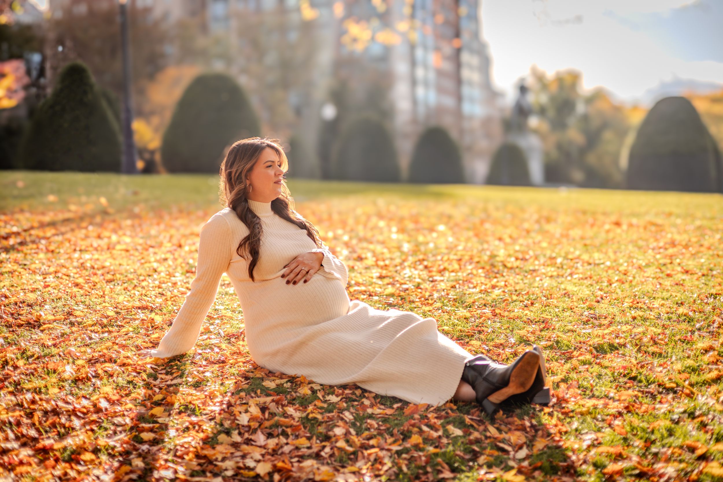 A pregnant woman in a white dress sits on golden autumn leaves during a sunny fall afternoon.