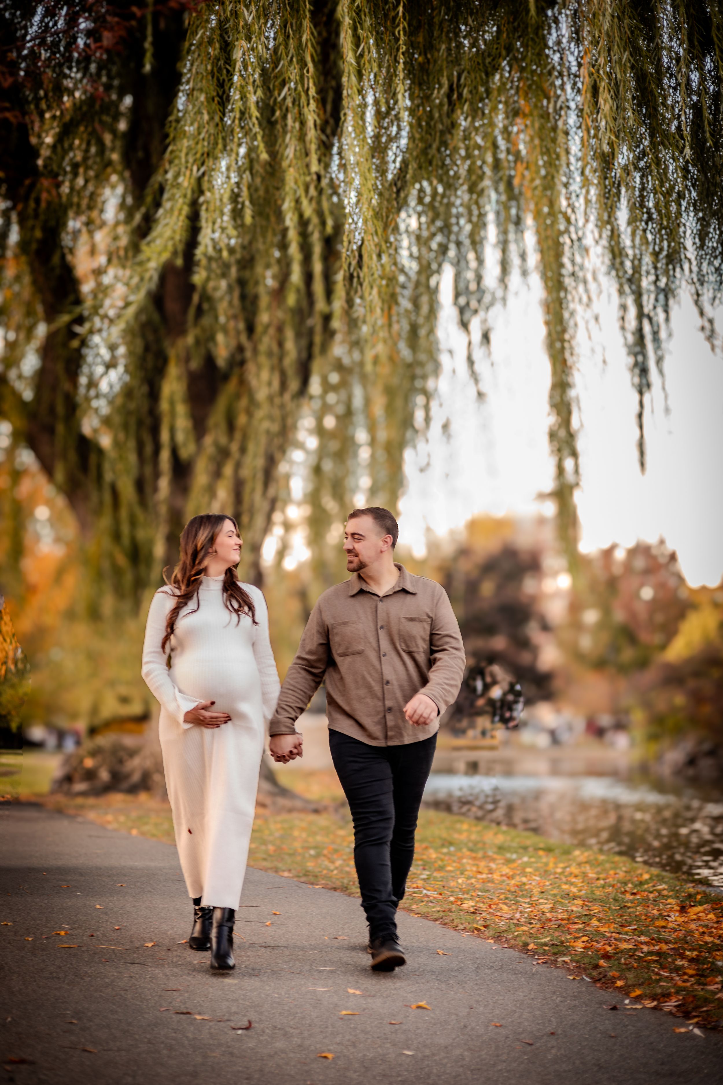 A couple walks hand-in-hand along a pathway under weeping willow trees on an autumn afternoon.
