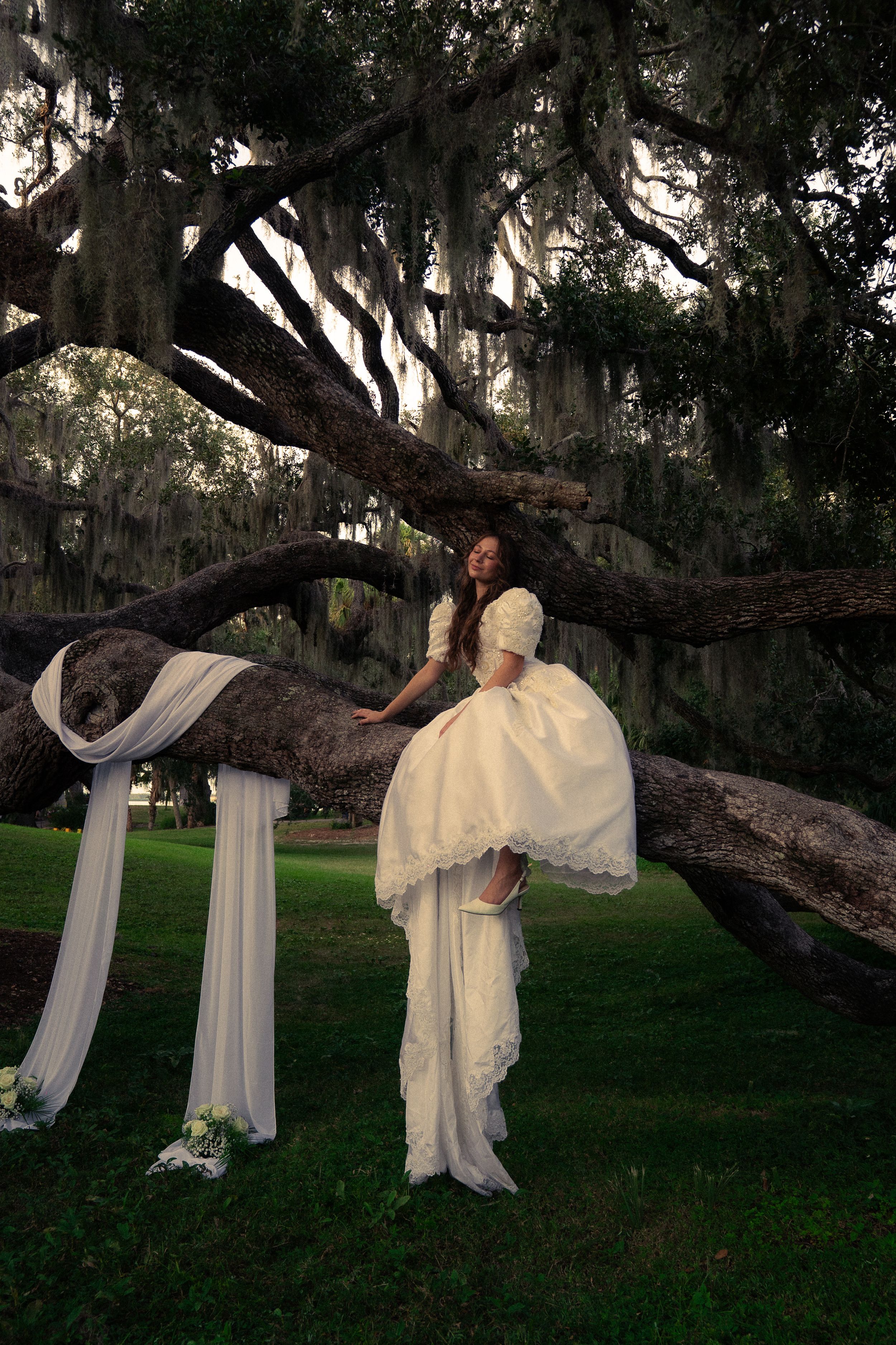 A wedding dress with a long flowing veil decorated with pink floral petals trailing along a sunlit wooden floor.