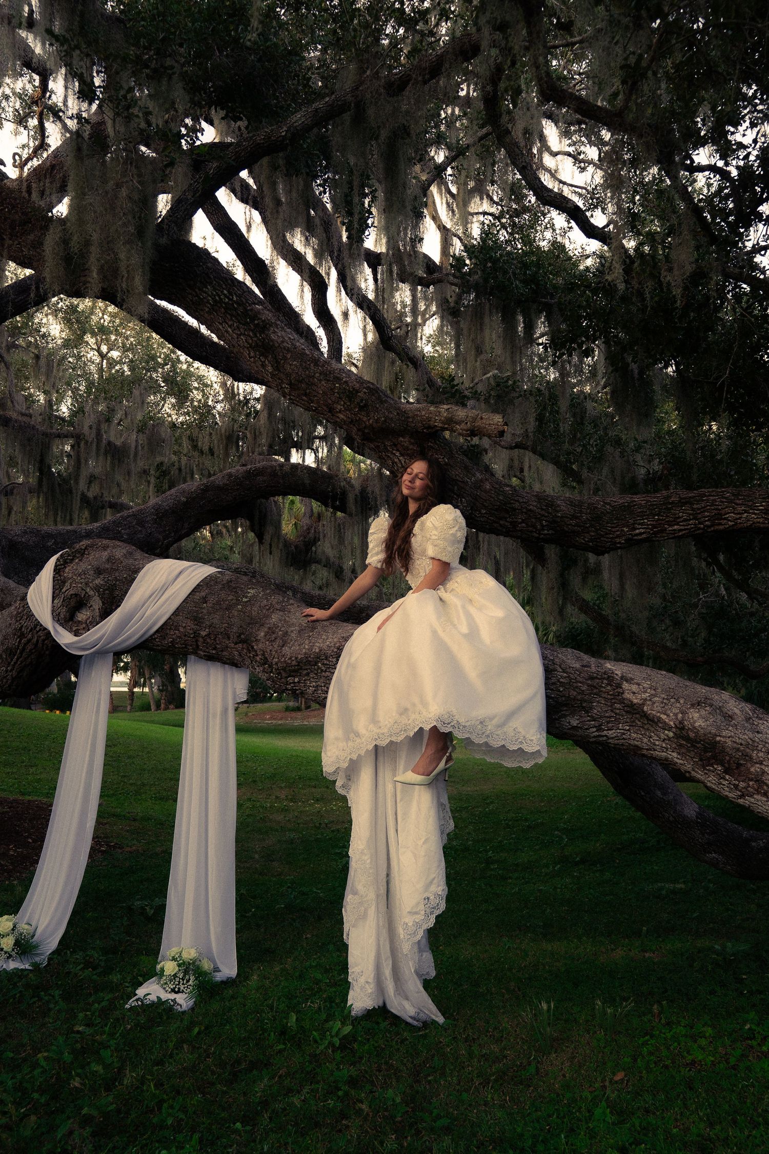 A wedding dress with a long flowing veil decorated with pink floral petals trailing along a sunlit wooden floor.