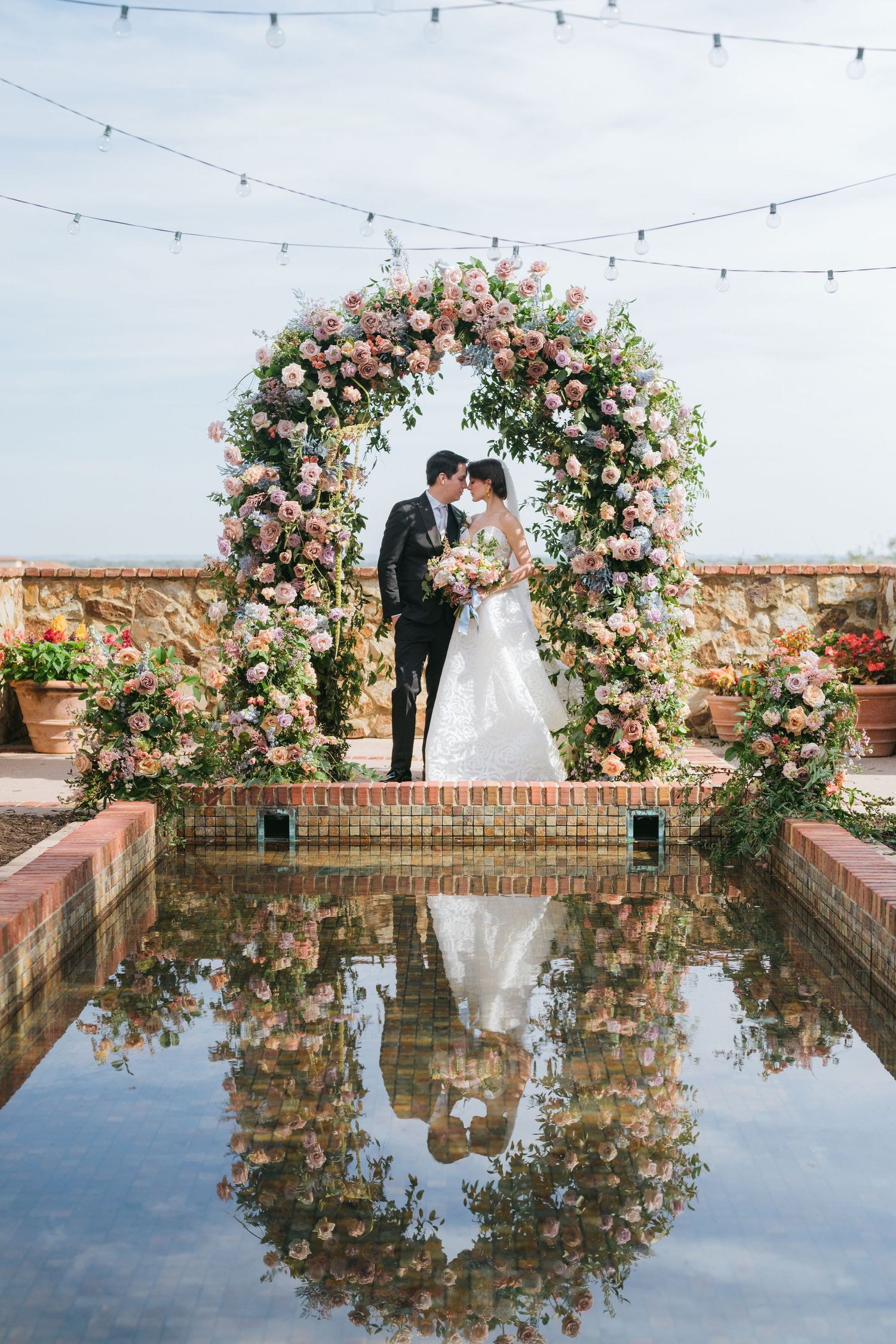 Romantic floral arch reflected in water with string lights creating a dreamy outdoor wedding setting.
