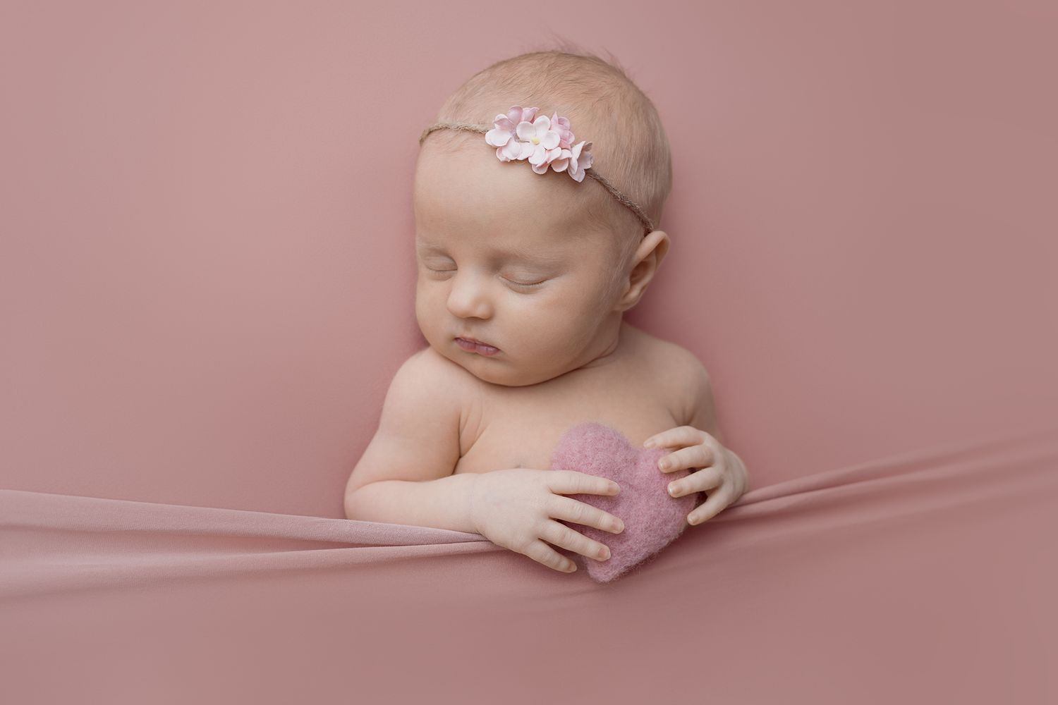 A sleeping baby wearing a delicate floral headband reclines on soft pink fabric with one tiny hand resting gently.