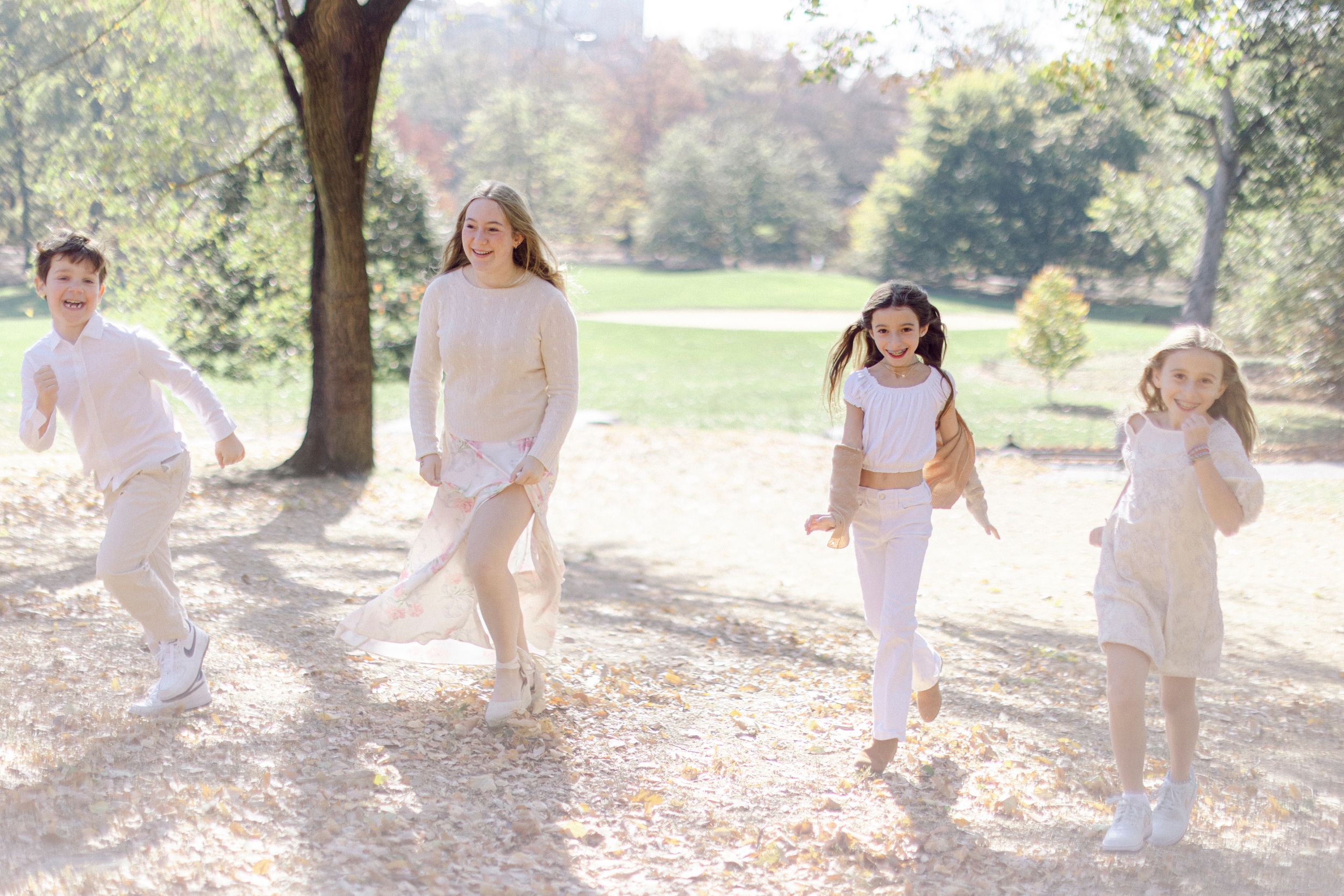 Four people in light colored clothing run playfully through a park on an autumn day.