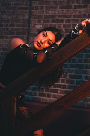 Artistic portrait against brick wall with dramatic lighting and wooden beams, styled in dark tones and orange accents.