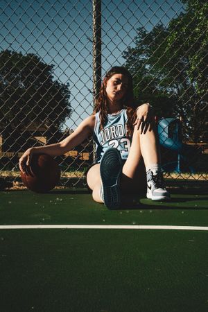 Basketball player in retro jersey sitting on outdoor court with chain link fence backdrop.